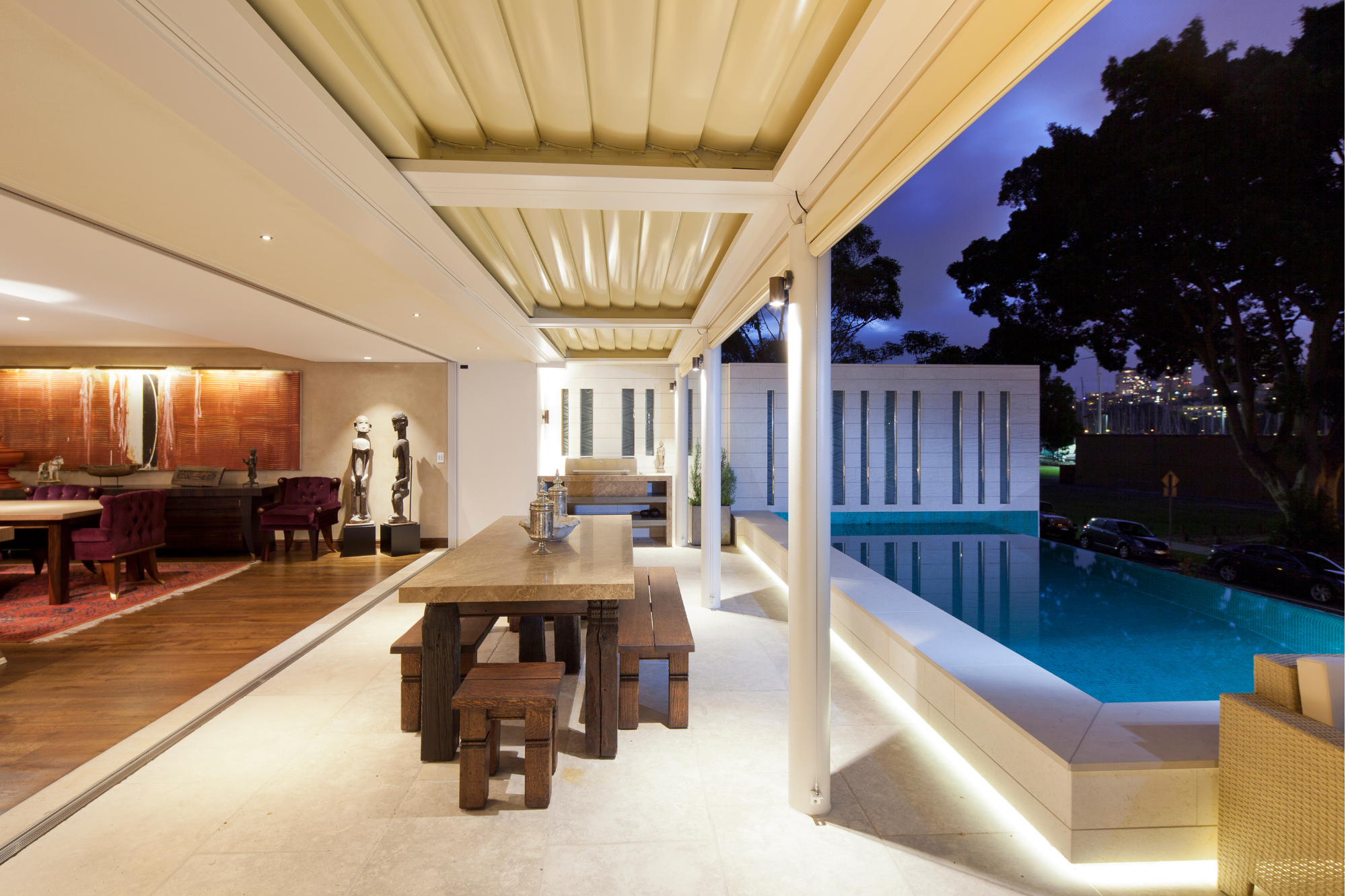 Modern outdoor patio and indoor living space at dusk, with a swimming pool, wooden dining table, all well lit with custom lighting. Contemporary art and sculpture in the background.
