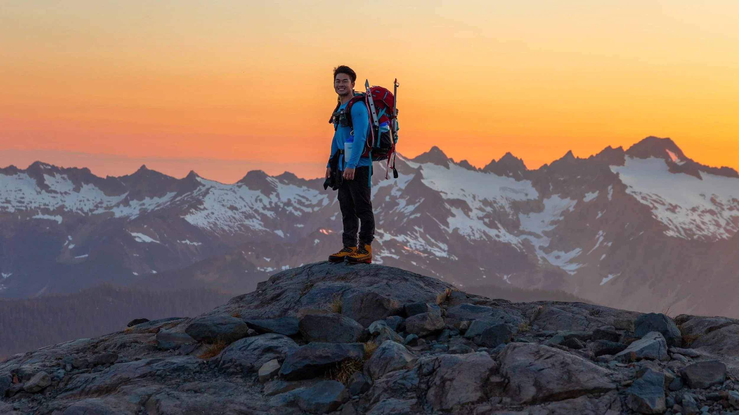 A male hiker wearing a blue jacket, black pants, and yellow hiking boots stands on rocks in a mountainous landscape during sunset, with snow-capped peaks in the background.