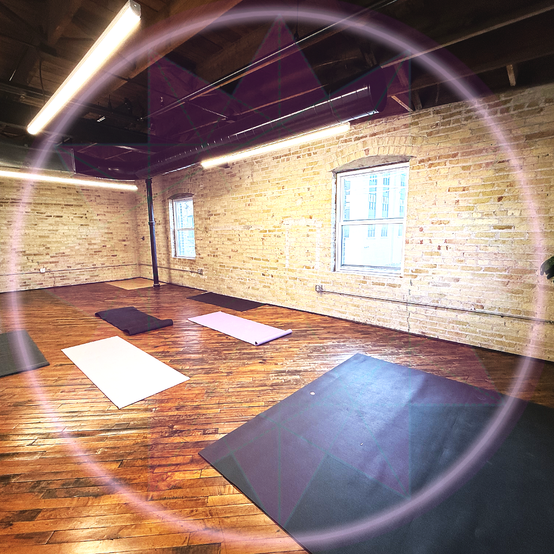 An empty yoga studio with hardwood floors, brick walls, large windows, and yoga mats arranged on the floor.