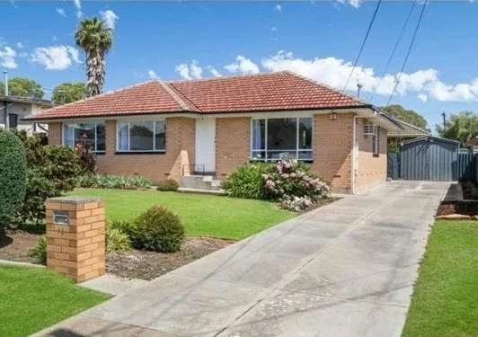 A single-story brick house with a red-tiled roof, large windows, a concrete driveway, front garden with bushes and flowers, and a blue garage in the backyard.