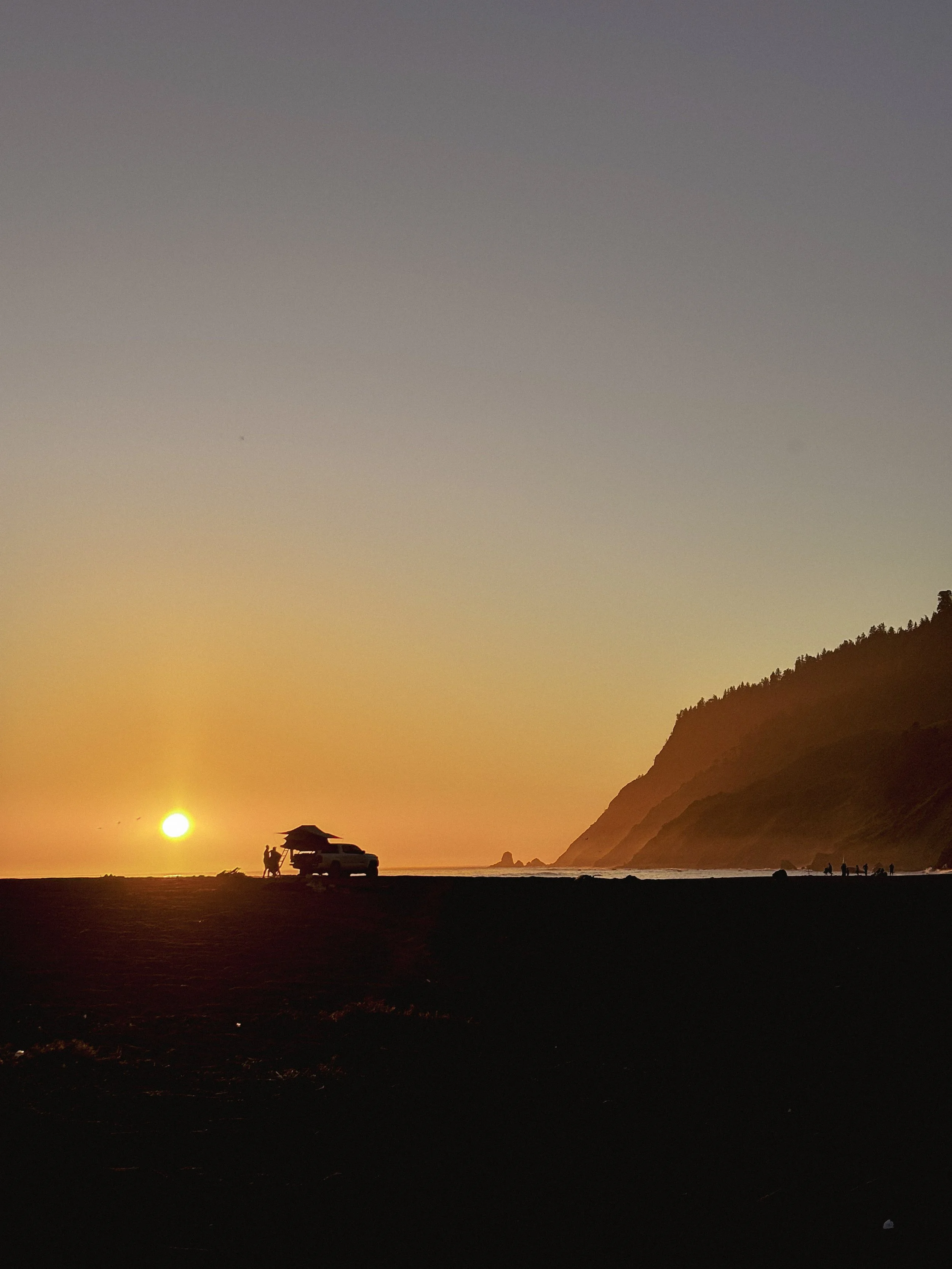Coucher de soleil sur une plage avec un véhicule et une tente, silhouettes de personnes, et des falaises en arrière-plan.
