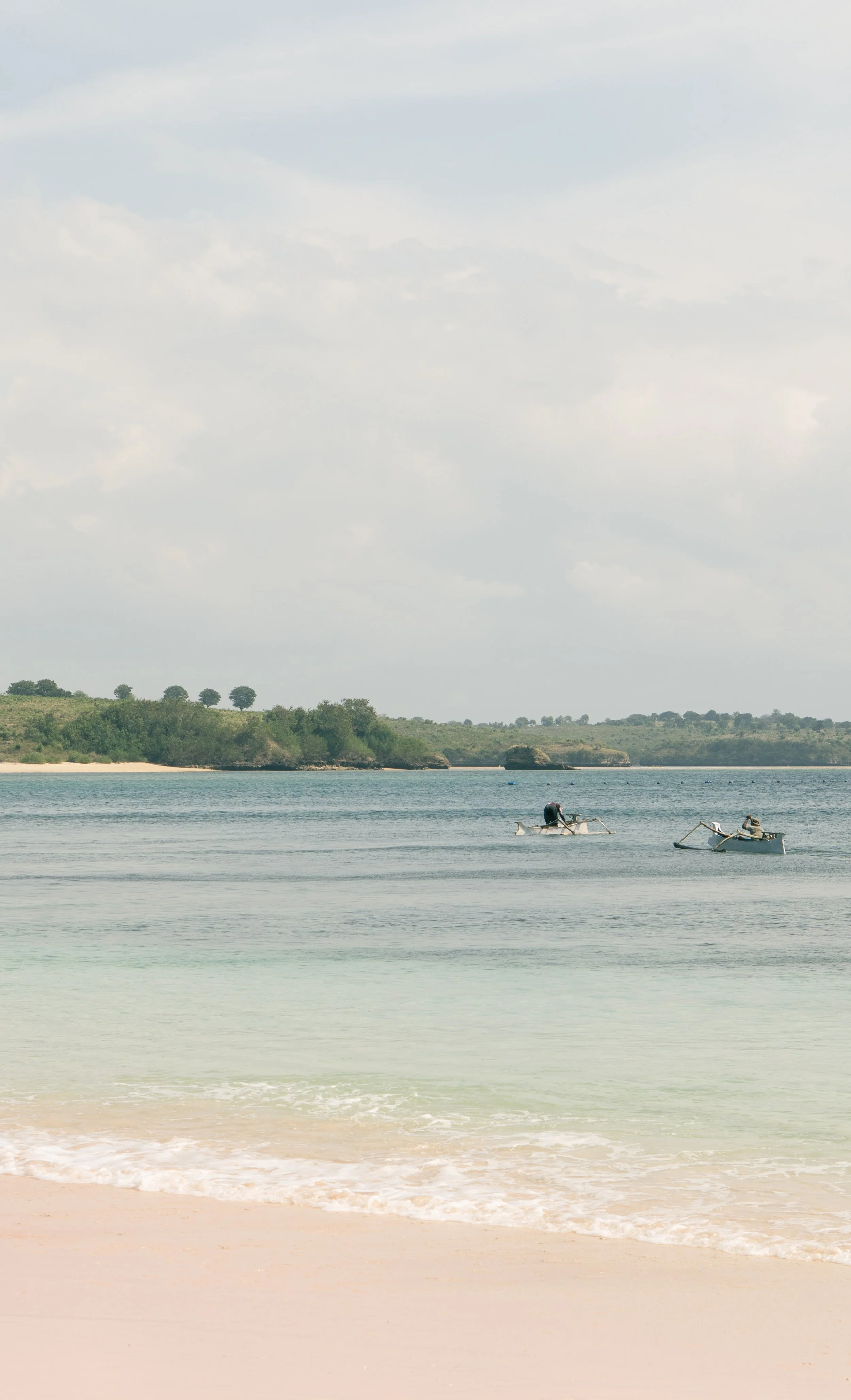 Plage avec deux petits bateaux à moteur sur l'eau, côté sable, avec une côte verdoyante en arrière-plan et un ciel partiellement nuageux.