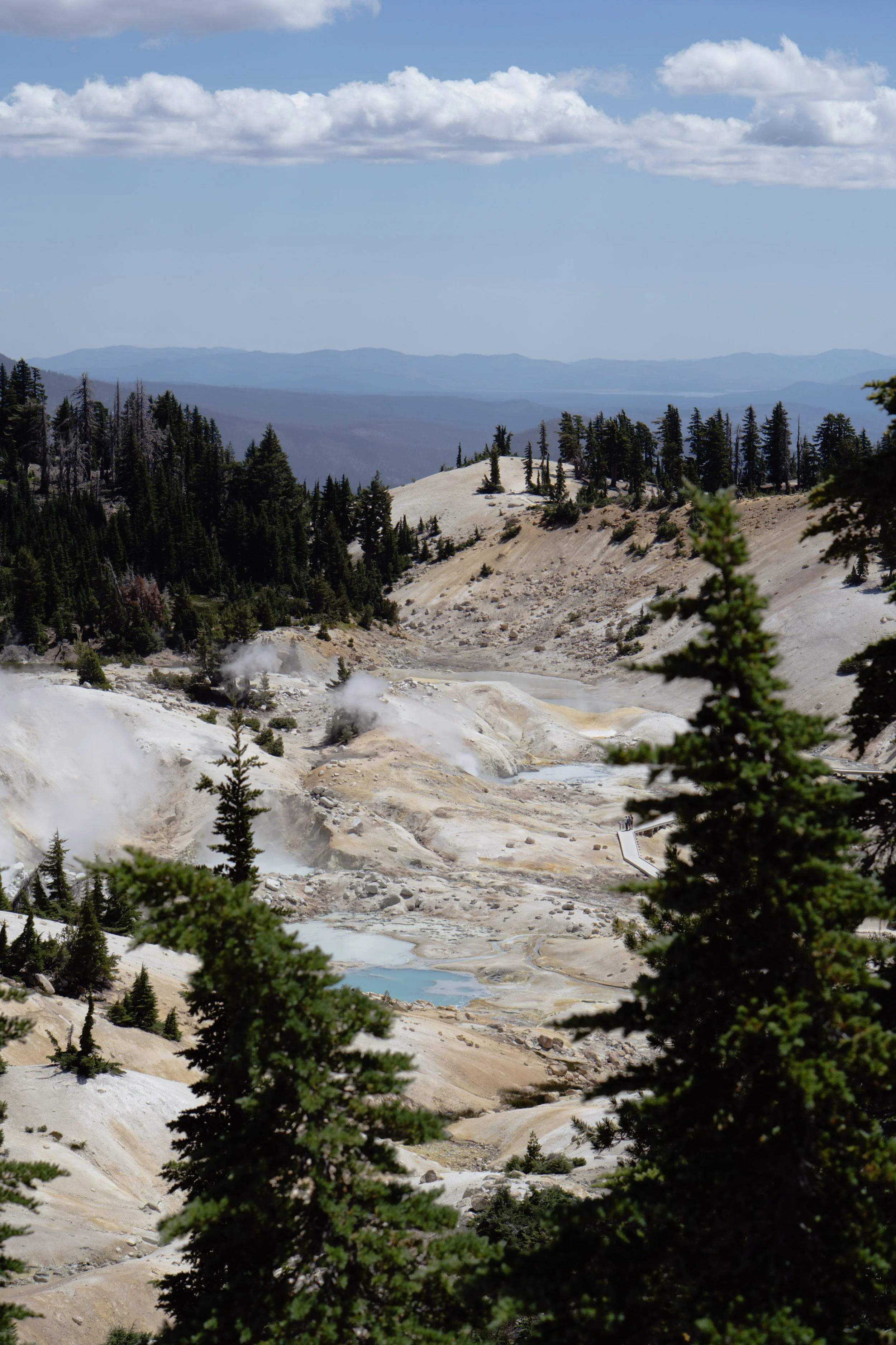 Paysage de sources thermales dans un parc naturel avec des montagnes en arrière-plan, des arbres verts et un ciel partiellement nuageux.