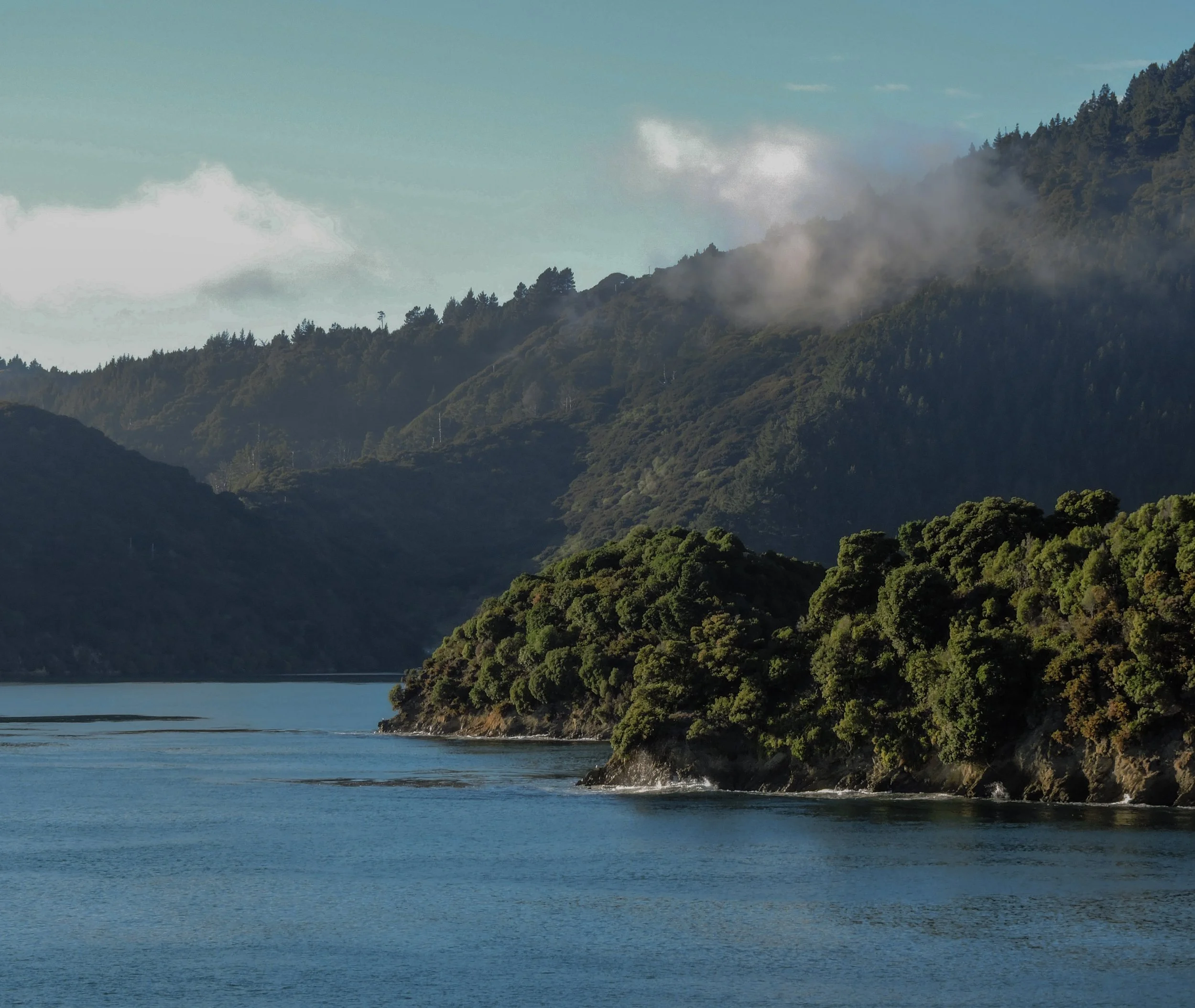 Paysage de montagnes verdoyantes et rivière calmi avec des nuages entourant les sommets.