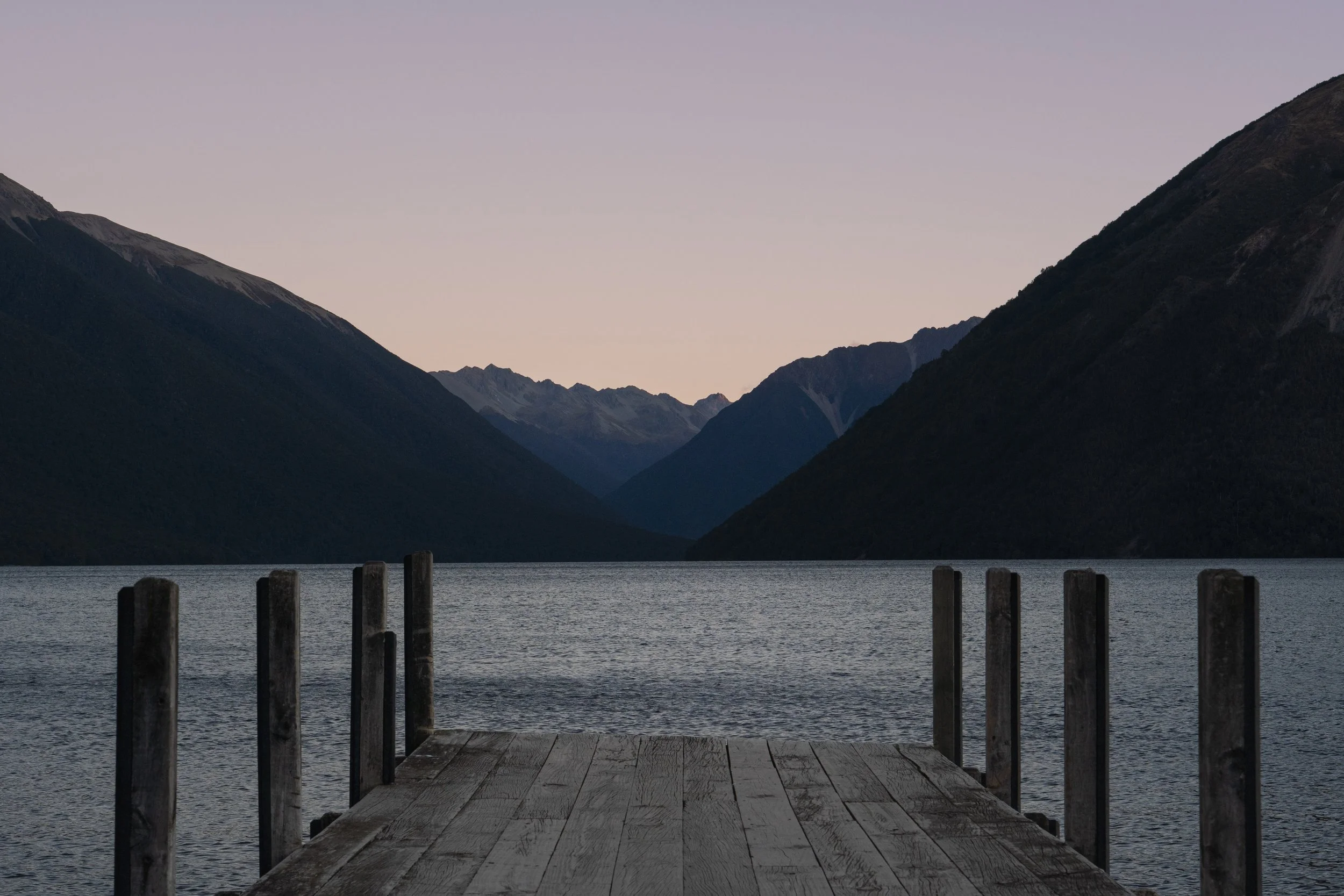 Un pont en bois donnant sur un lac entouré de montagnes, au crépuscule.