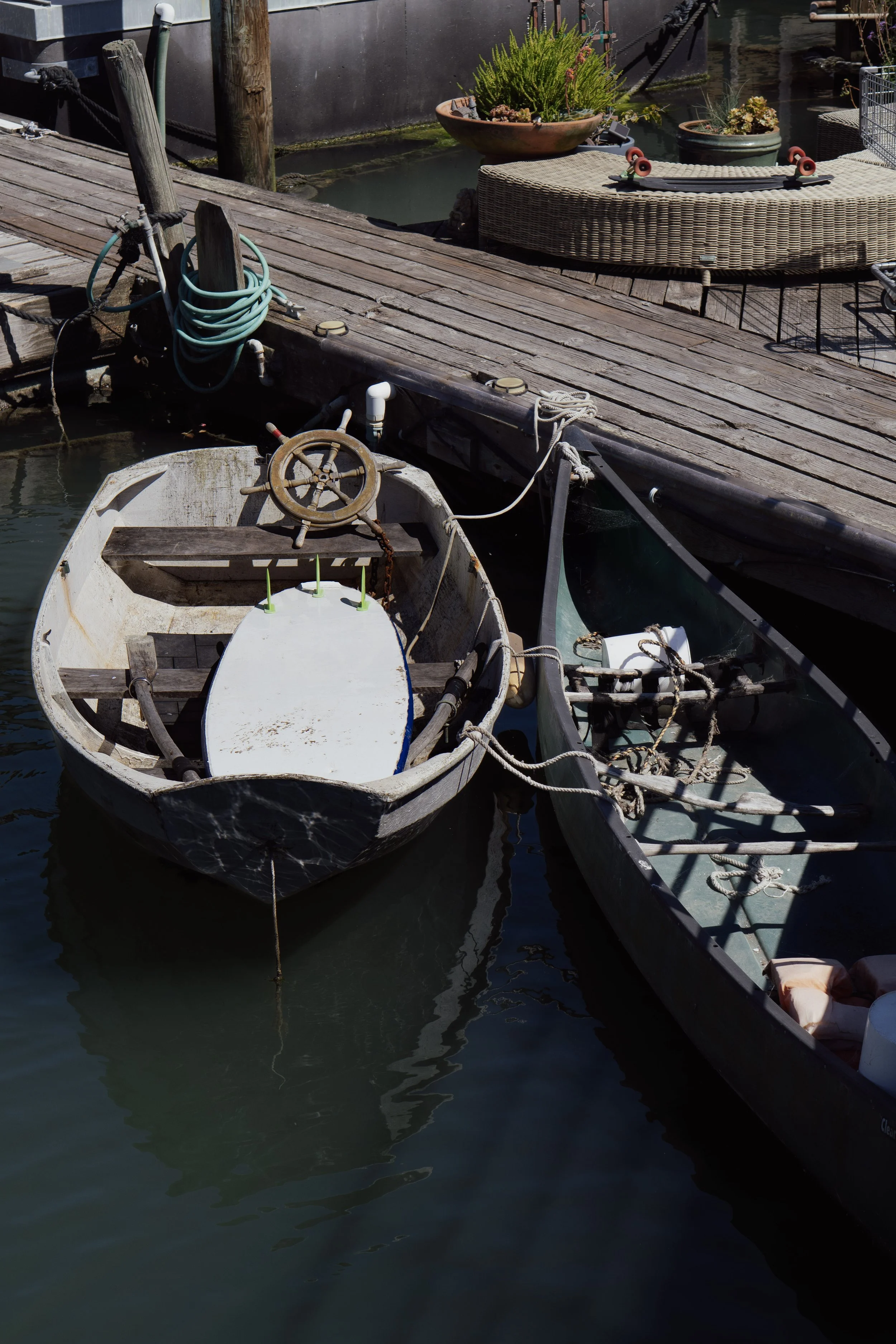 Deux bateaux en bois amarrés à un pont en bois, avec des cordages et un gouvernail, près d'une terrasse avec des plantes et une roue de skate.