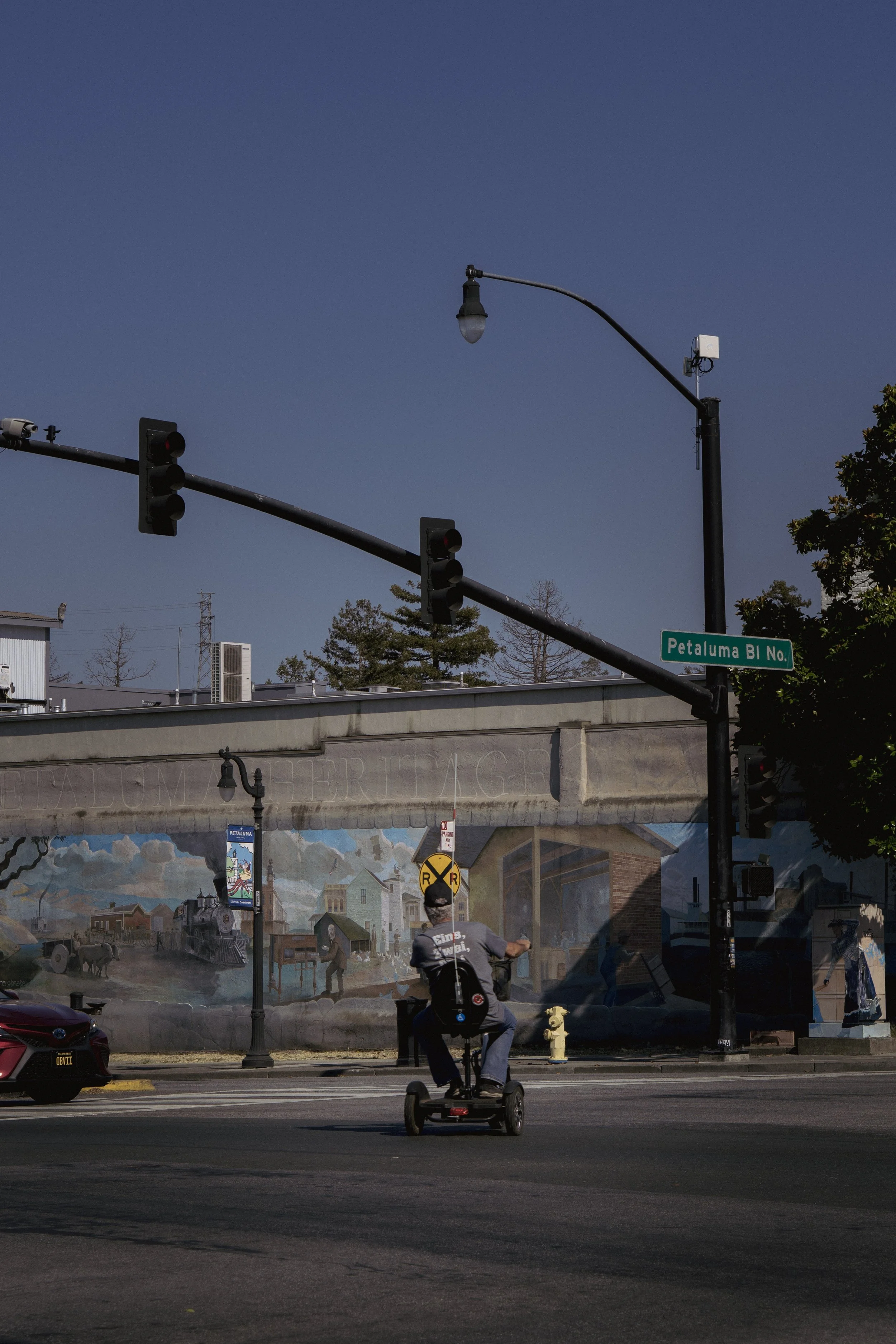 Un homme en fauteuil électrique traverse une intersection, avec des feux de circulation et une signalisation routière, une peinture murale en arrière-plan et un arbre à droite.