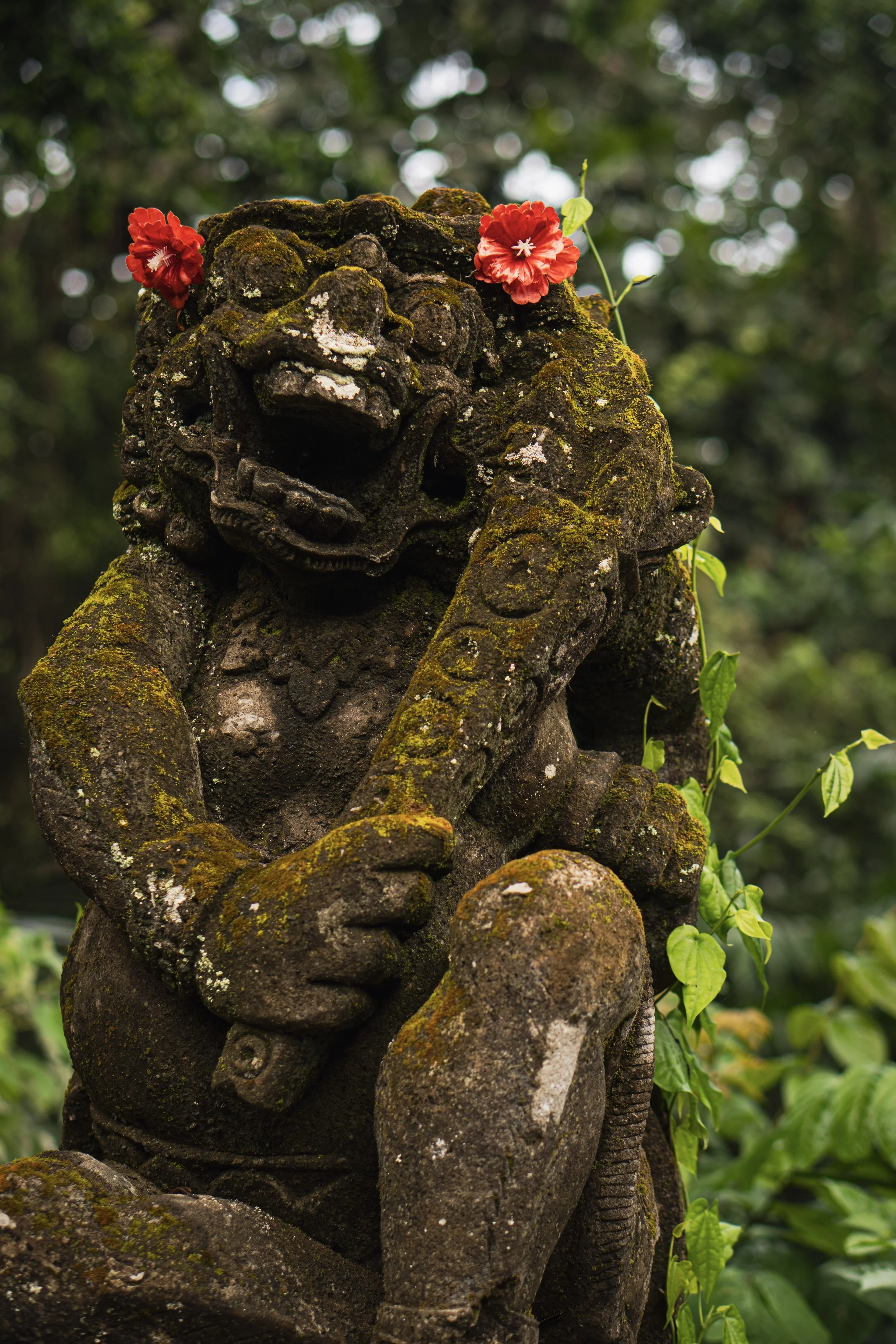 Statue en pierre de un lion avec des fleurs rouges sur la tête, dans un environnement naturel verdoyant.