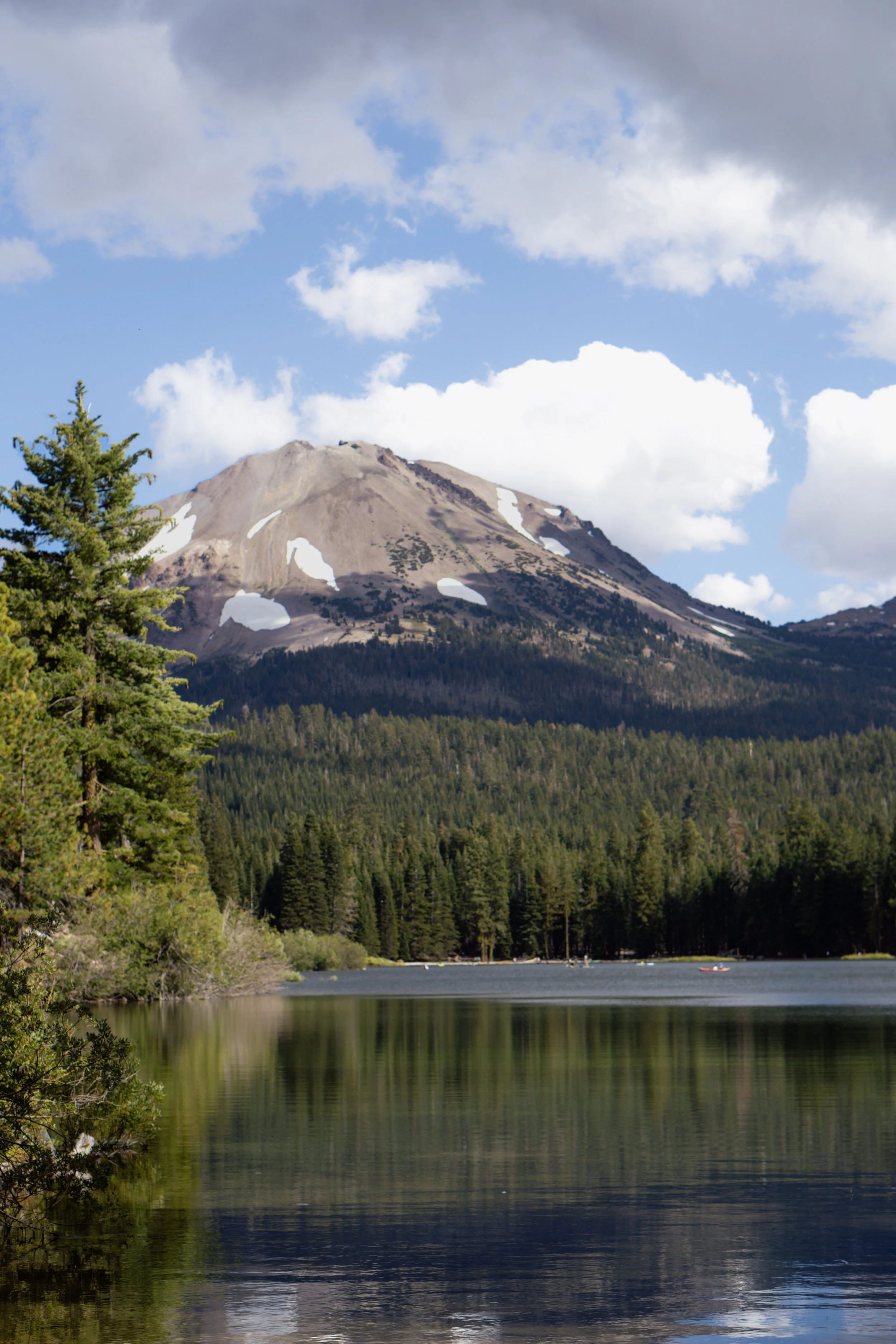 Montagne avec sommets enneigés entourée de forêt et un lac au premier plan, ciel partiellement nuageux.