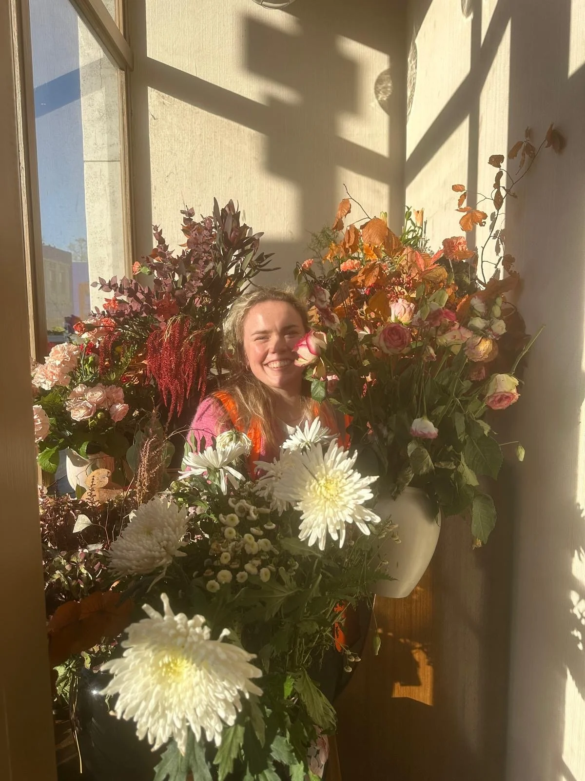 A woman with long hair smiling behind a large arrangement of colorful flowers near a window with sunlight streaming in.