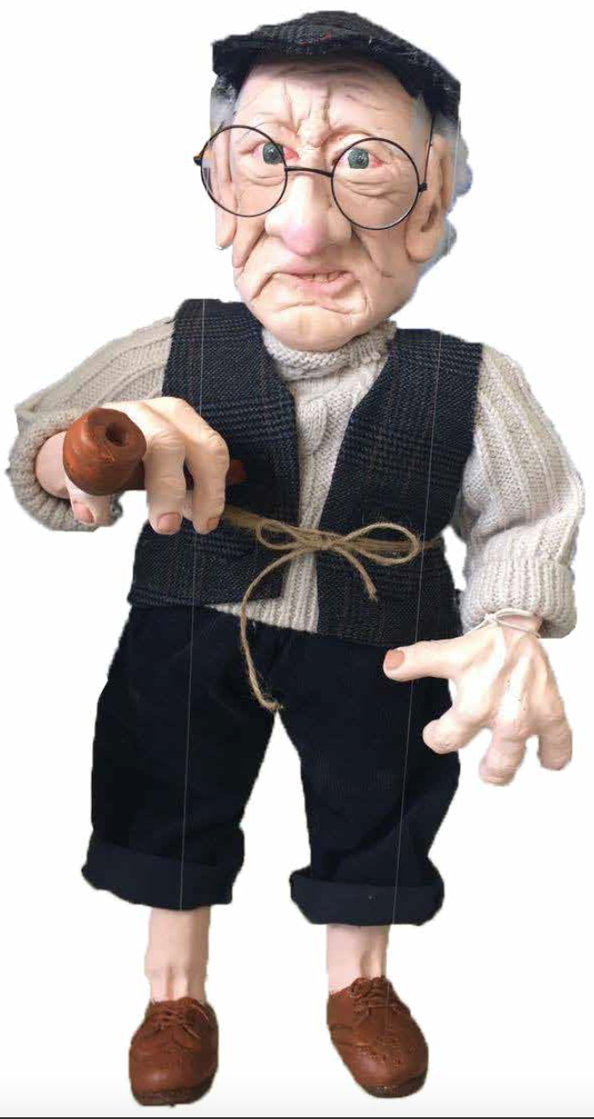 A puppet with exaggerated, wrinkled elderly face, wearing glasses, a black hat, a beige sweater, black vest, and black pants. Holding a pipe in one hand, with a string tied around the waist.
