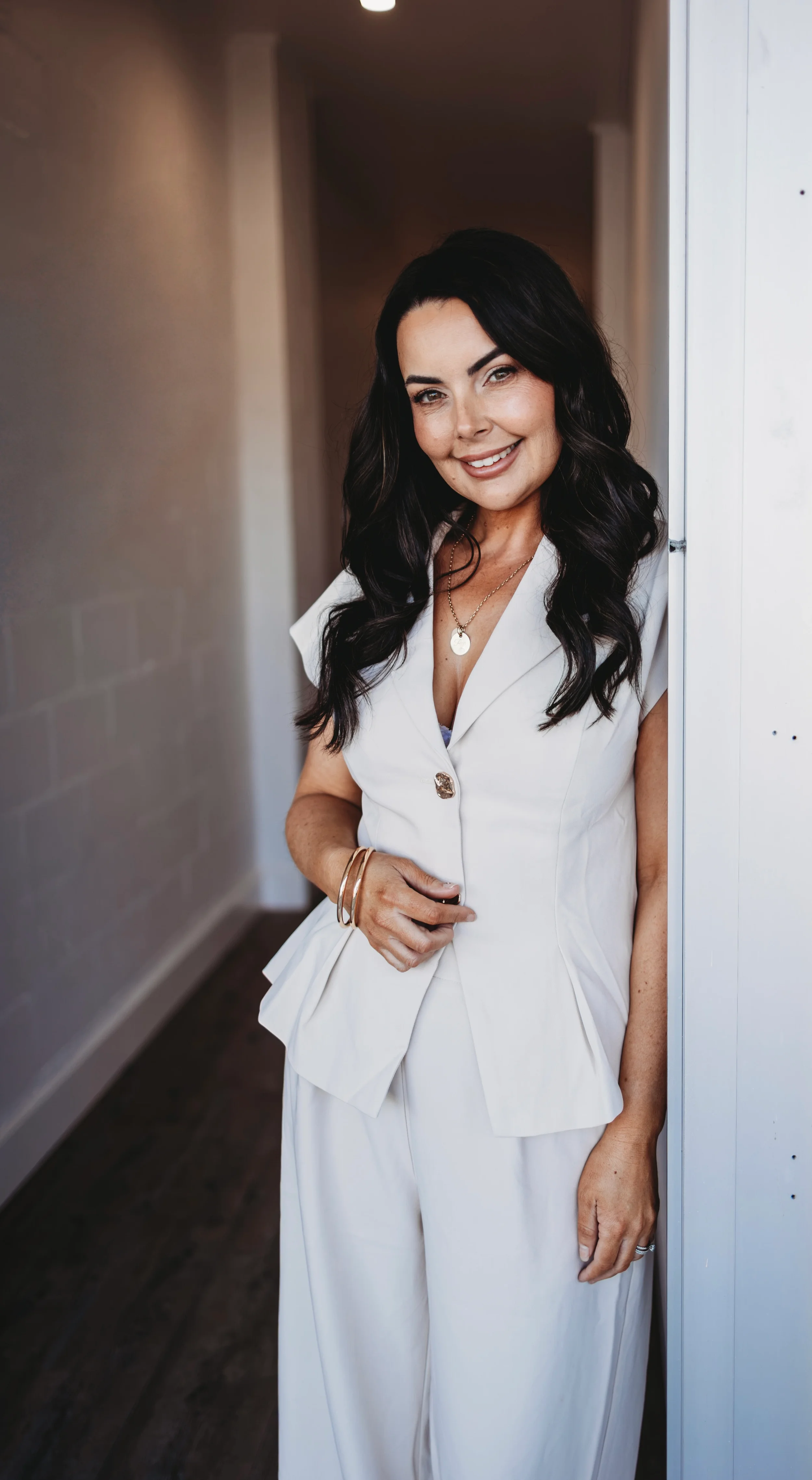 A woman with dark, wavy hair wearing a white suit and jewelry, standing in a hallway and smiling at the camera.