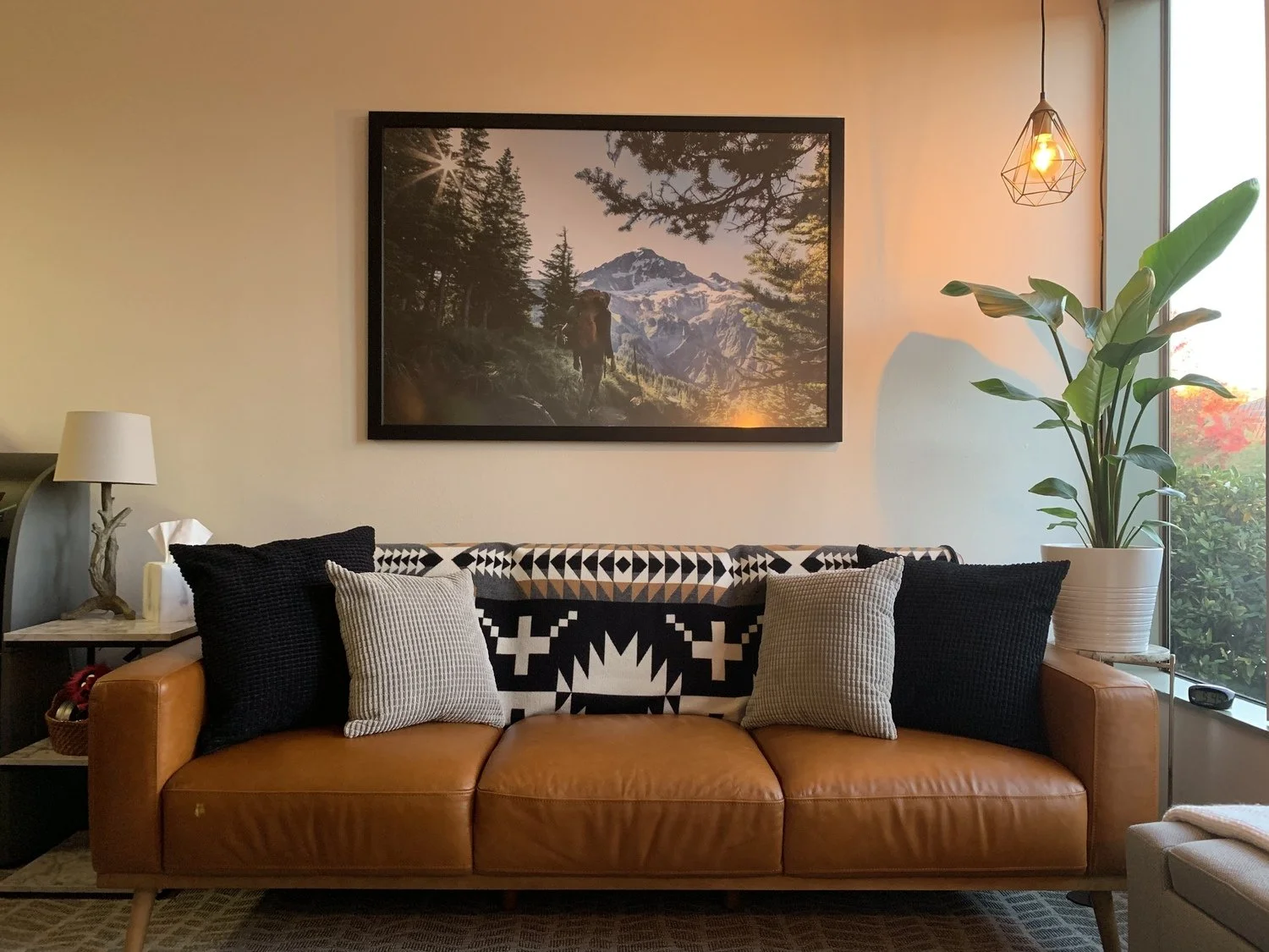 Therapist office with a brown leather sofa, five decorative pillows, a side table with a lamp, a framed nature photograph, and a large potted plant near a window.