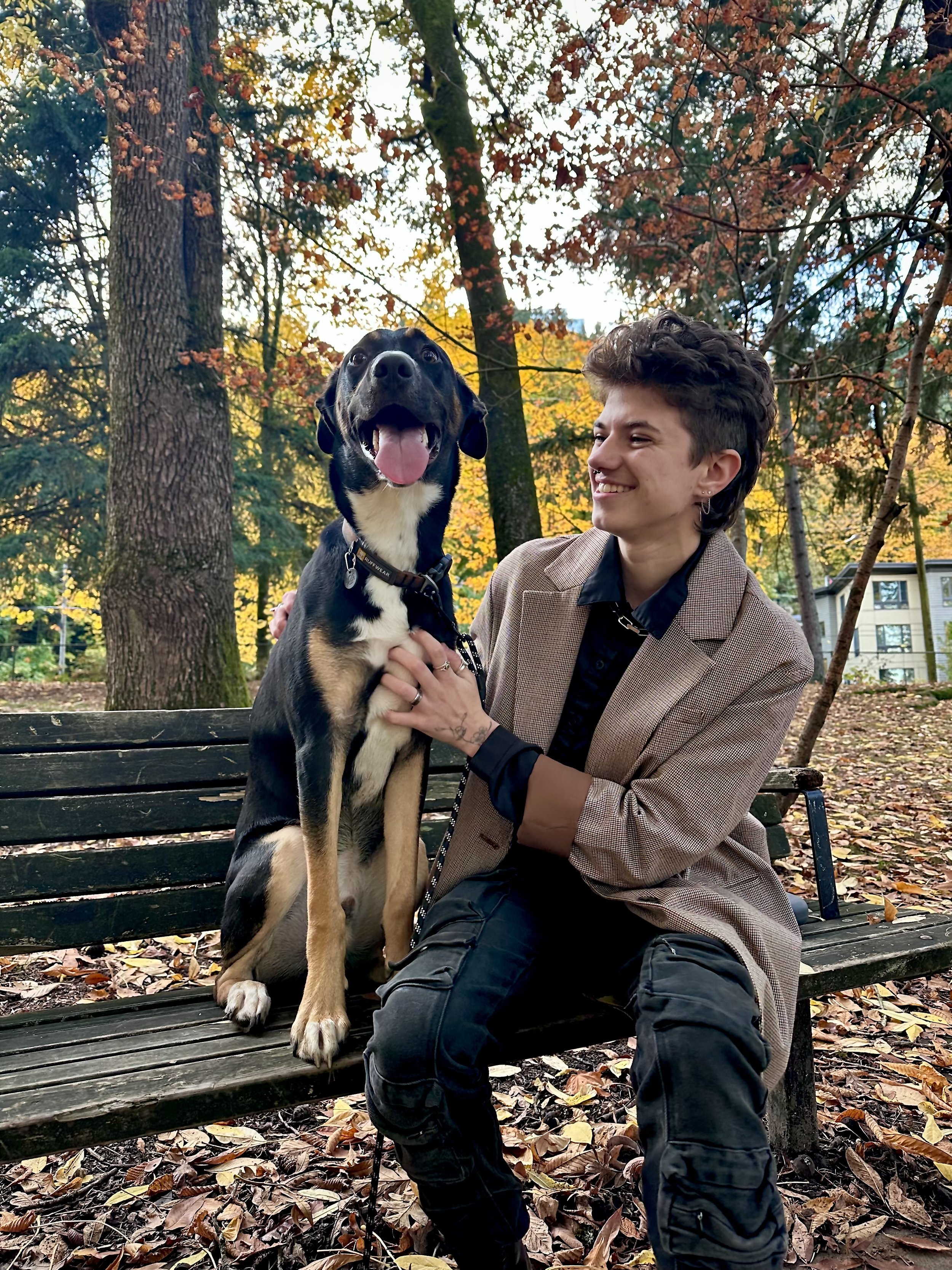 Person with short hair and earrings sitting on a bench outdoors, holding a large black and tan dog with its tongue out, during autumn in a park with fall leaves on the ground.