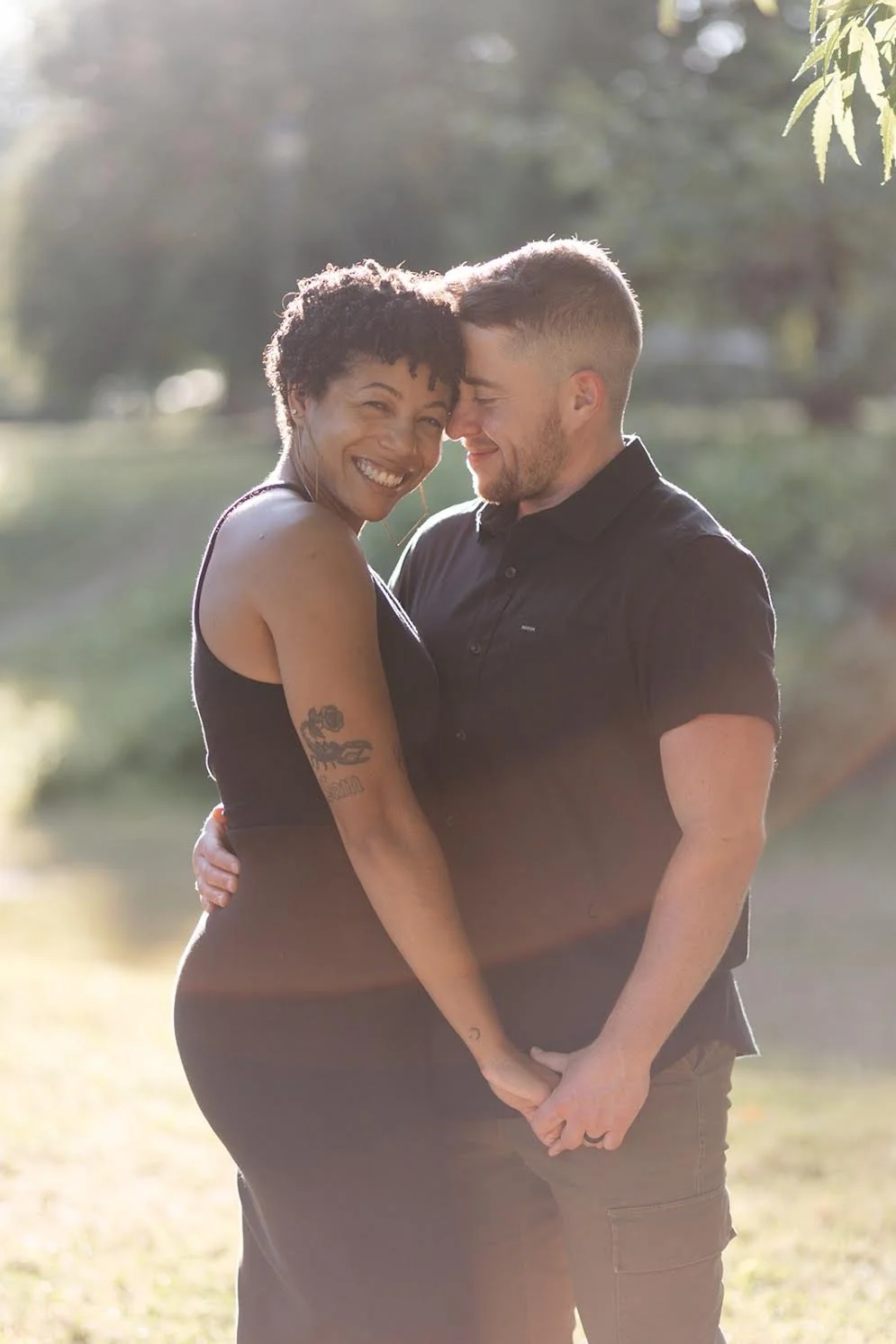 A smiling couple standing closely outdoors, holding hands and touching foreheads with sunlight streaming behind them.