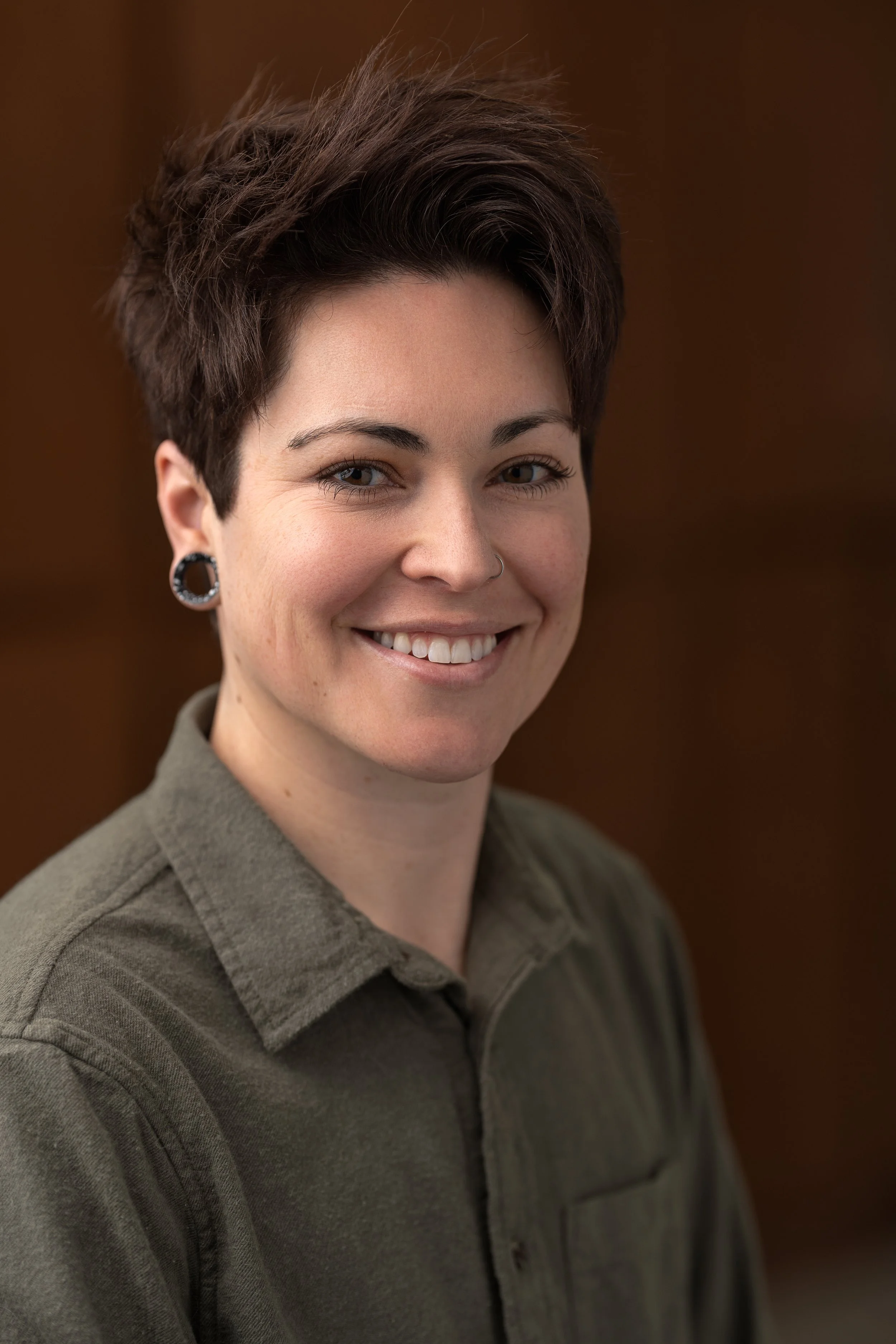 Headshot of a nonbinary therapist with short dark hair, wearing a green shirt, smiling, and a nose ring, against a reddish-brown background.
