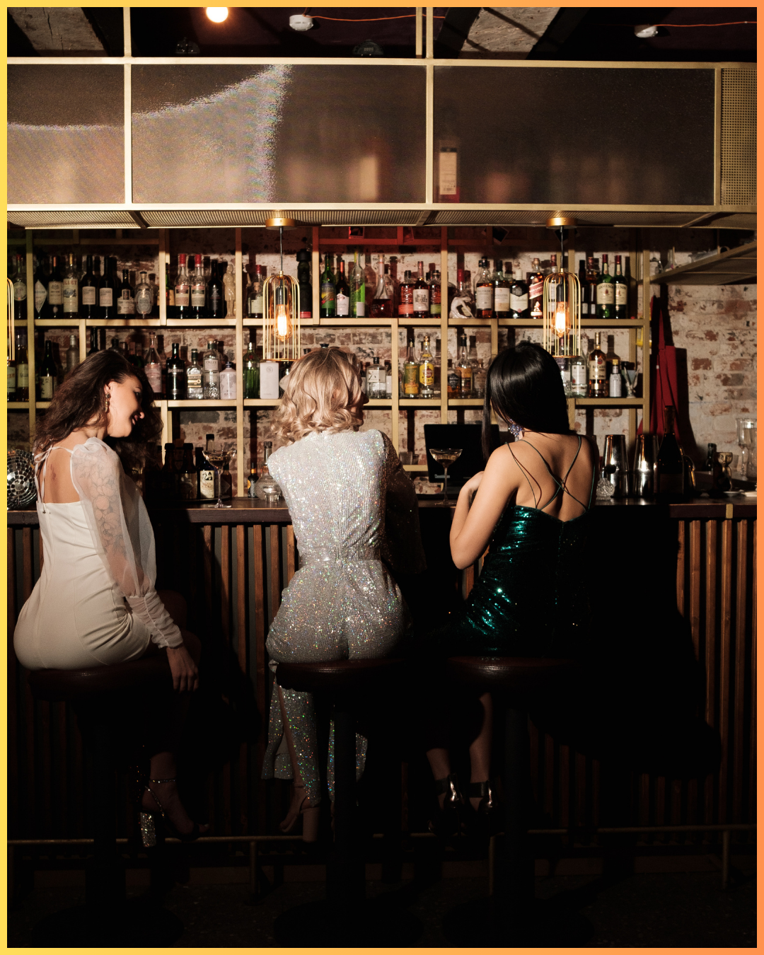 Three women sitting at a bar in a dimly lit pub, wearing glamorous dresses, with a background of shelves filled with liquor bottles and warm lighting.