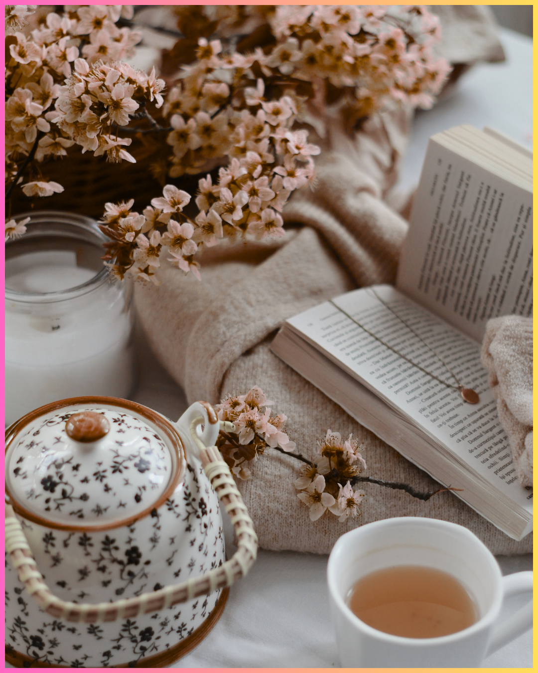 A cozy scene with pink cherry blossoms, a teapot, a cup of tea, a candle, an open book, and a blanket all arranged on a soft surface.