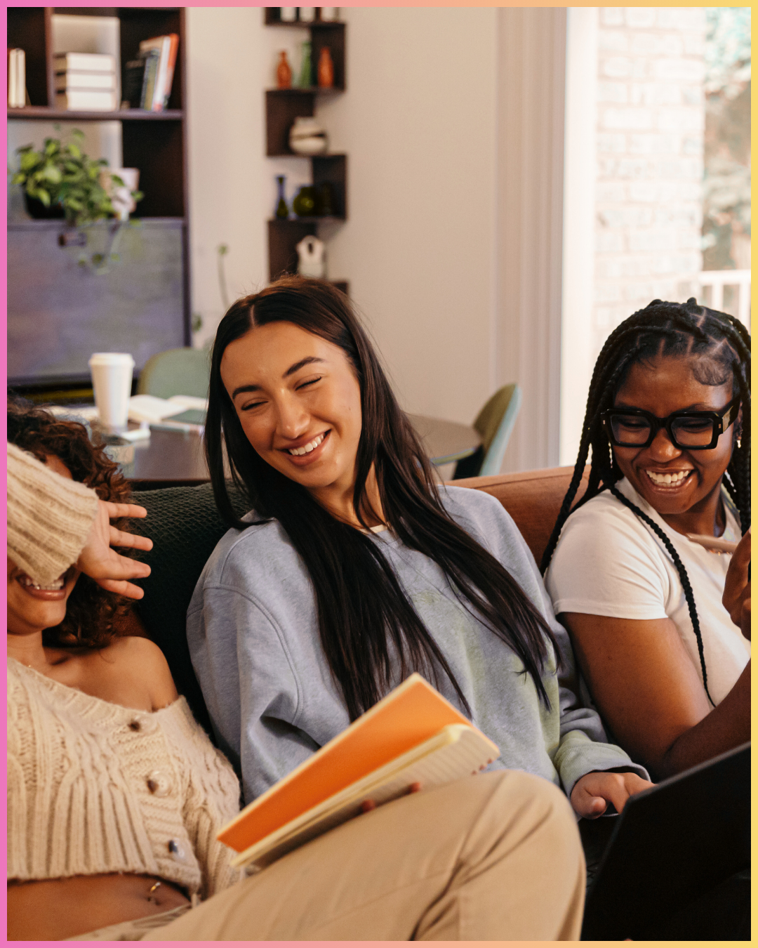 Three women sitting on a couch smiling and laughing, one holding a notebook. They are in a cozy living room with a bookshelf, a dining table in the background, and a large window letting in natural light.