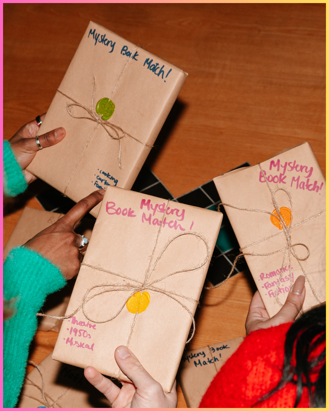 People holding wrapped mystery books with handwritten titles and colorful wax seals, on a wooden surface.