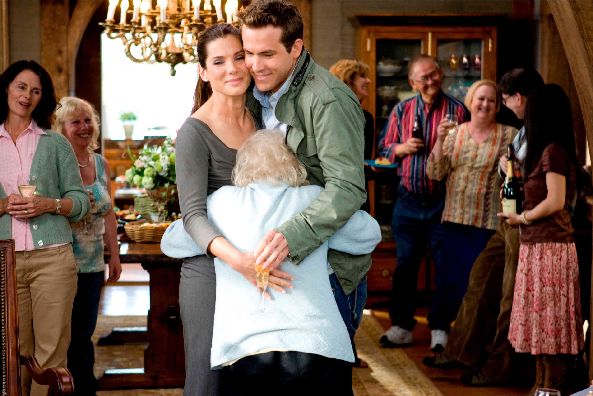 Scene from the  with Sandra Bollock &  Ryan Reynolds A family gathering in a cozy living room with people smiling and celebrating, including an elderly woman hugging a young woman and a young man, with others holding drinks in the background.