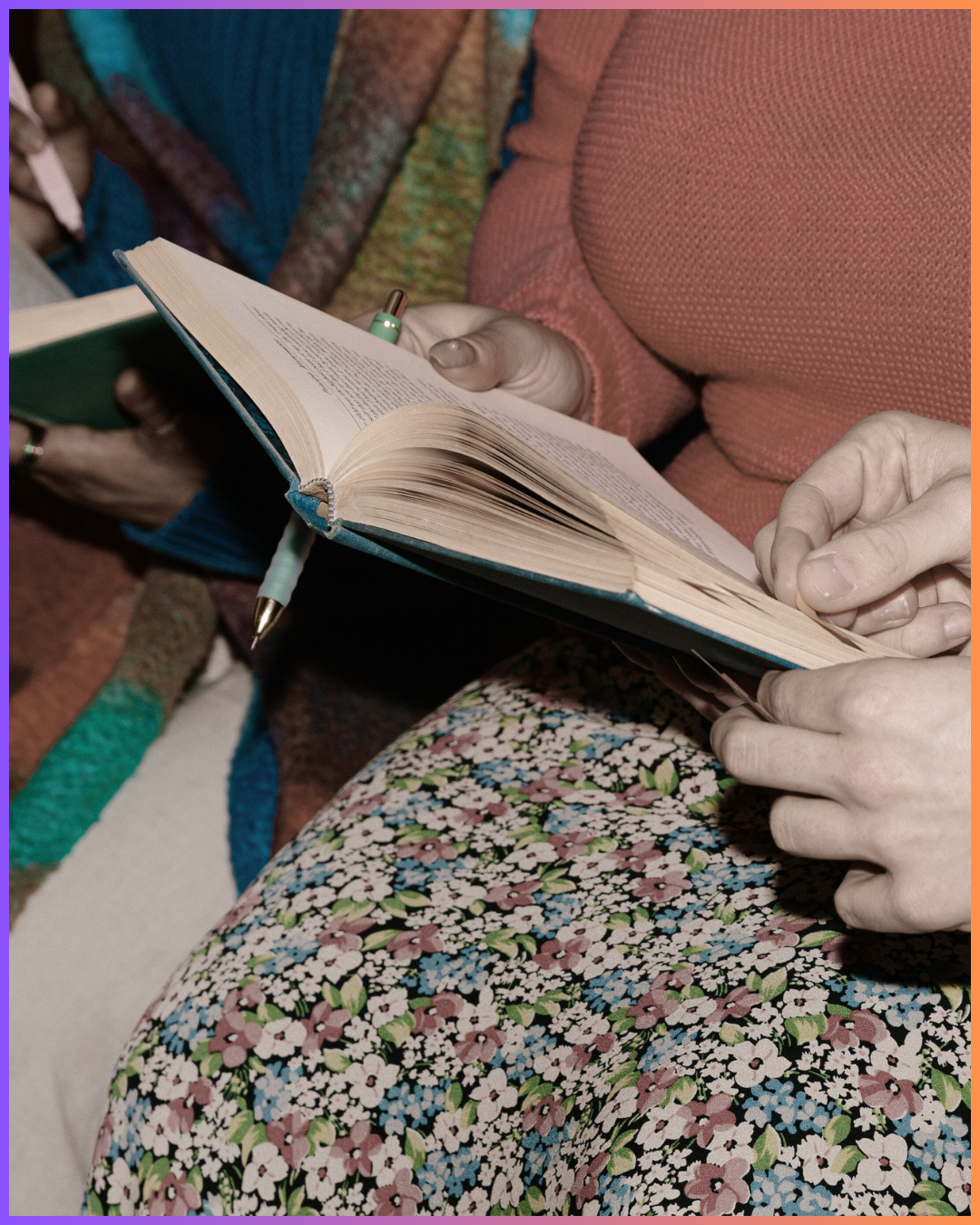 Close-up of a person holding an open book, with a pen in hand, while sitting on a surface with a colorful floral skirt and surrounded by others reading books.
