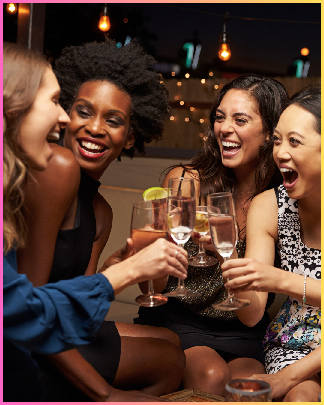 Four women sitting together at a bar, laughing and holding glasses of wine or cocktails, celebrating in a lively, decorated venue with warm lighting.