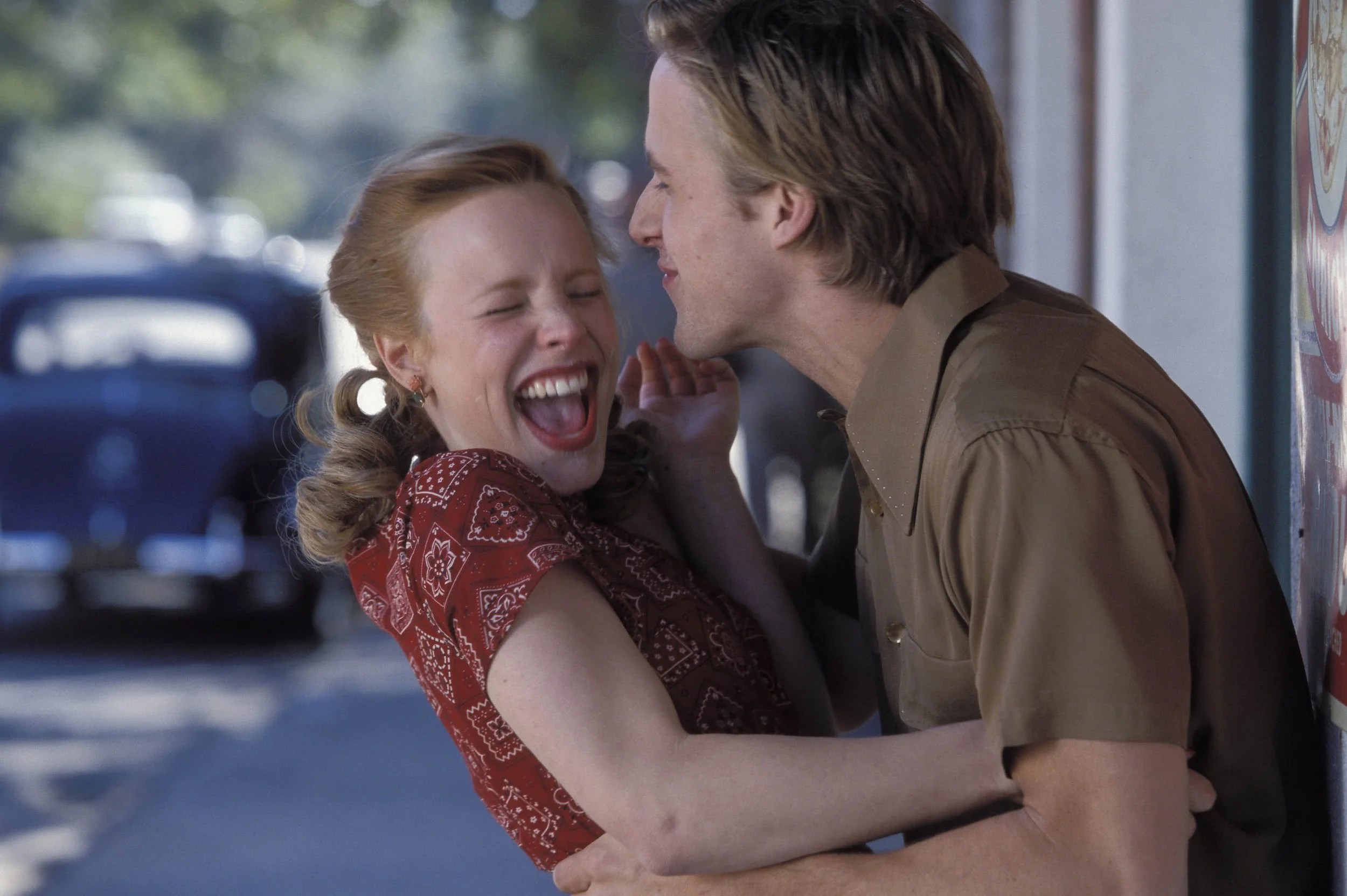 A scene from The notebook A young woman and a man are laughing and playing together on a sidewalk, with the man leaning in towards the woman and the woman smiling widely with her eyes closed.