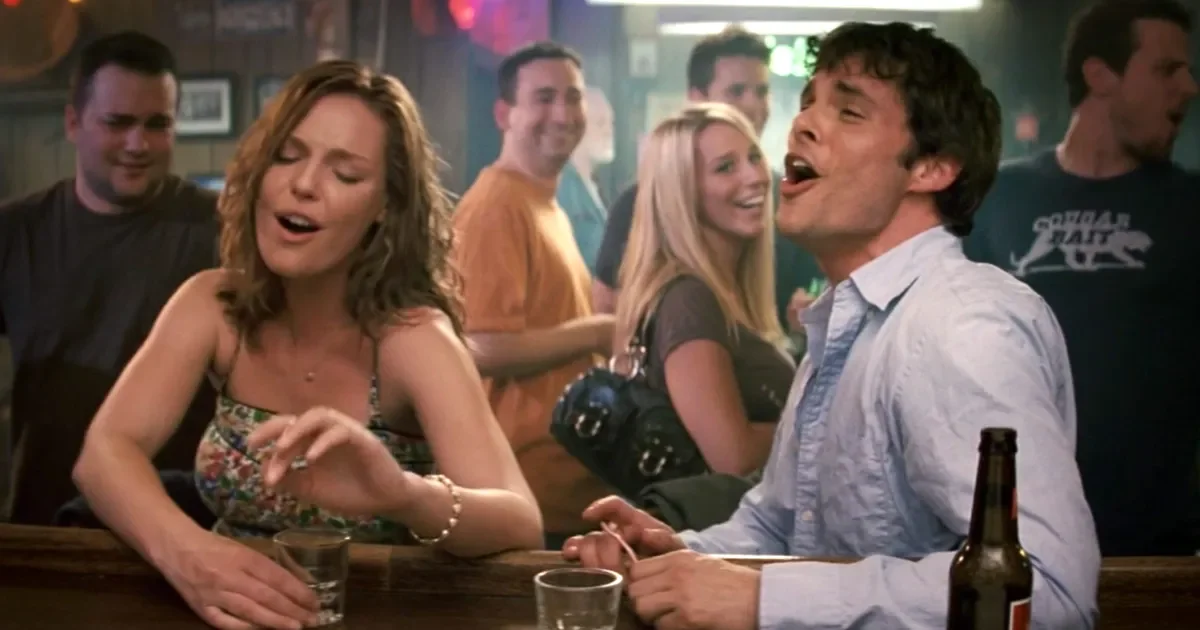 Katherine Heigl and  James Marsden in 27 Dresses singing and enjoying drinks at a bar with a couple in the foreground and others in the background smiling.