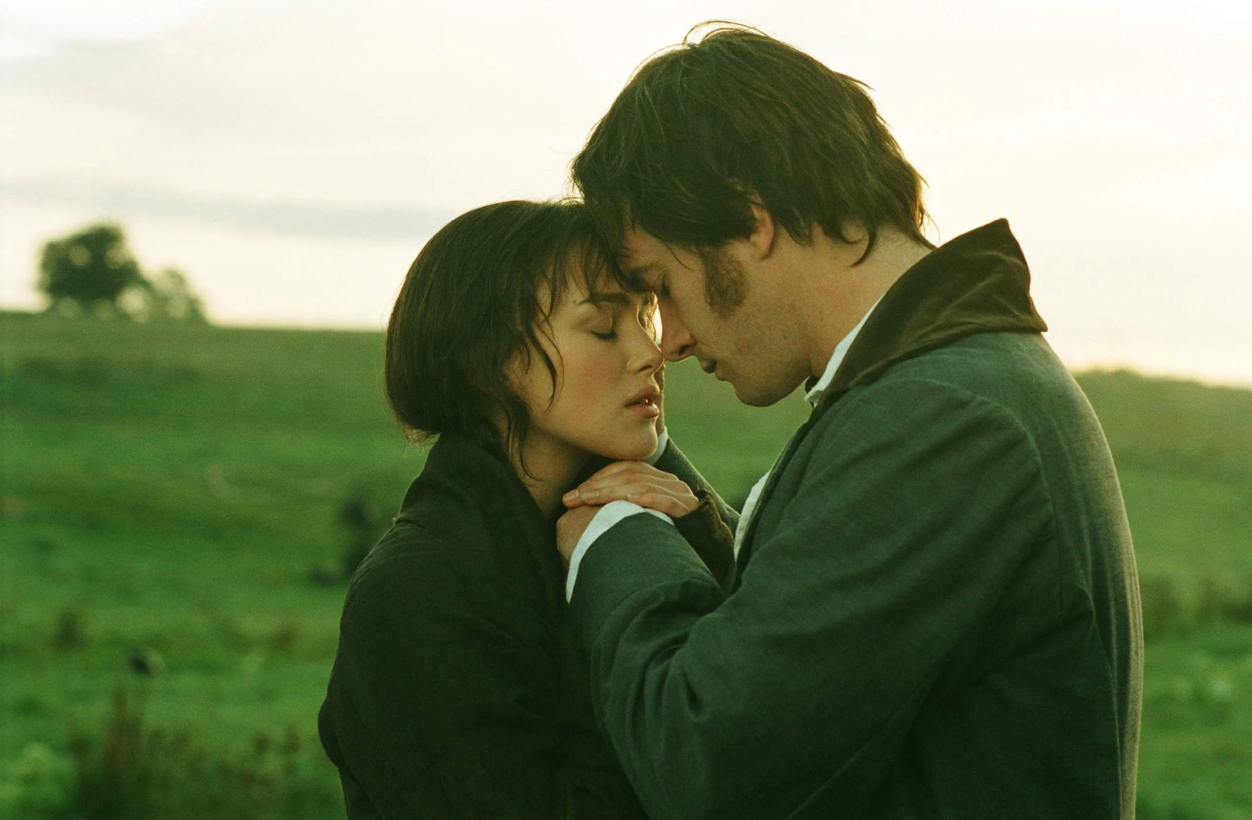 A Scene from Pride & Predujoice with Elizabeth & Mr. Darcy A man and woman are in a close embrace outdoors during sunset, with their foreheads touching and eyes closed, in a green field with blurred trees in the background.