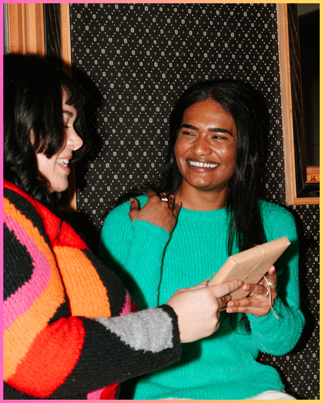 Two women smiling and looking at a phone, one wearing a colorful striped sweater, the other in a turquoise sweater, indoors with a dotted black and white wallpaper background.