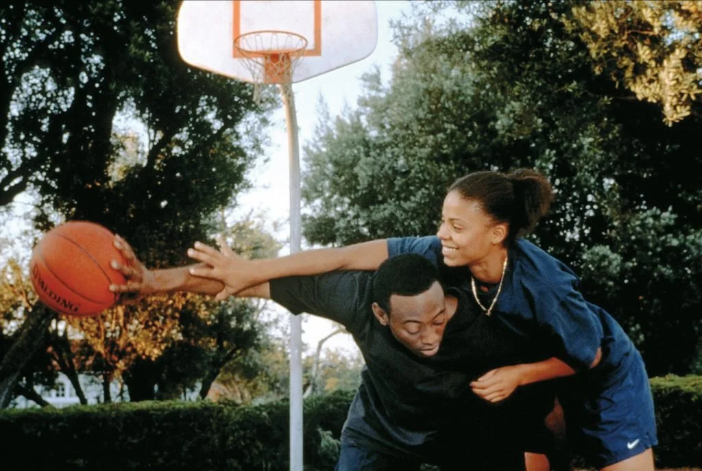 Scene from Love & basketball outdoors, one person with a ponytail is reaching for the ball while the other is bent over with their head down