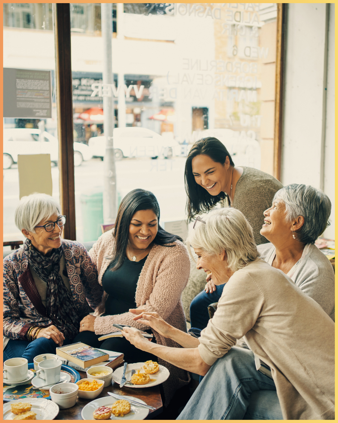 Five women happily gathered at a table in a cafe, looking at a smartphone together and smiling, with cups and snacks on the table.