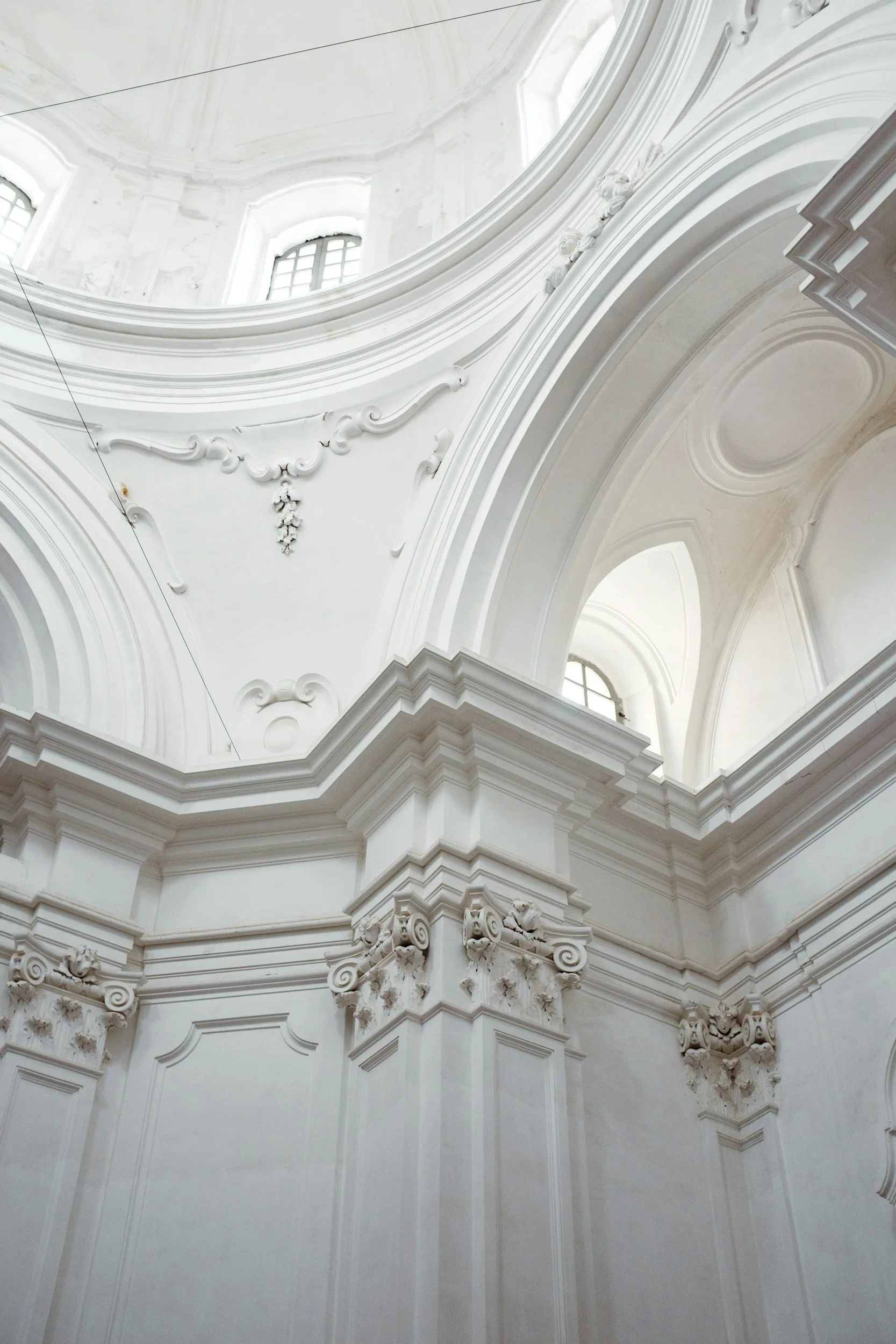 Interior view of a white, ornate classical dome with detailed moldings and windows, conveying calm and refinement.