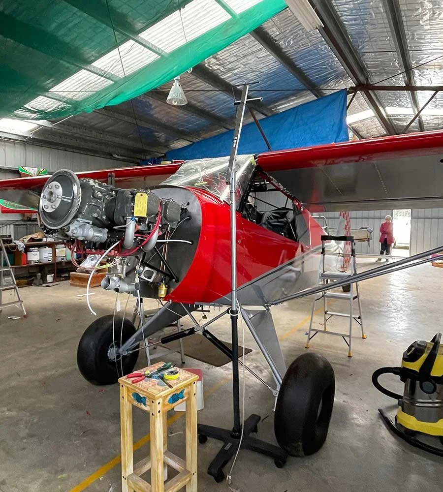 Small red and silver aircraft inside a hangar, with tools and equipment nearby, and a person in the background.