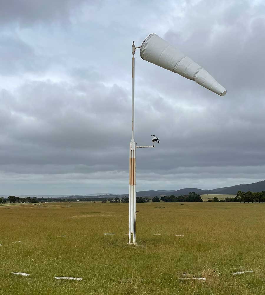A weather wind sock and wind vane mounted on a tall pole in an open field under a cloudy sky.
