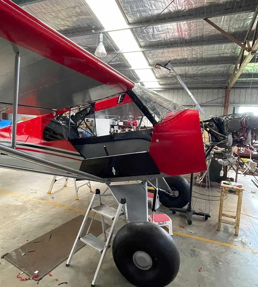 A small red and black aircraft inside a hangar, with a ladder and tools nearby, in a workshop setting.