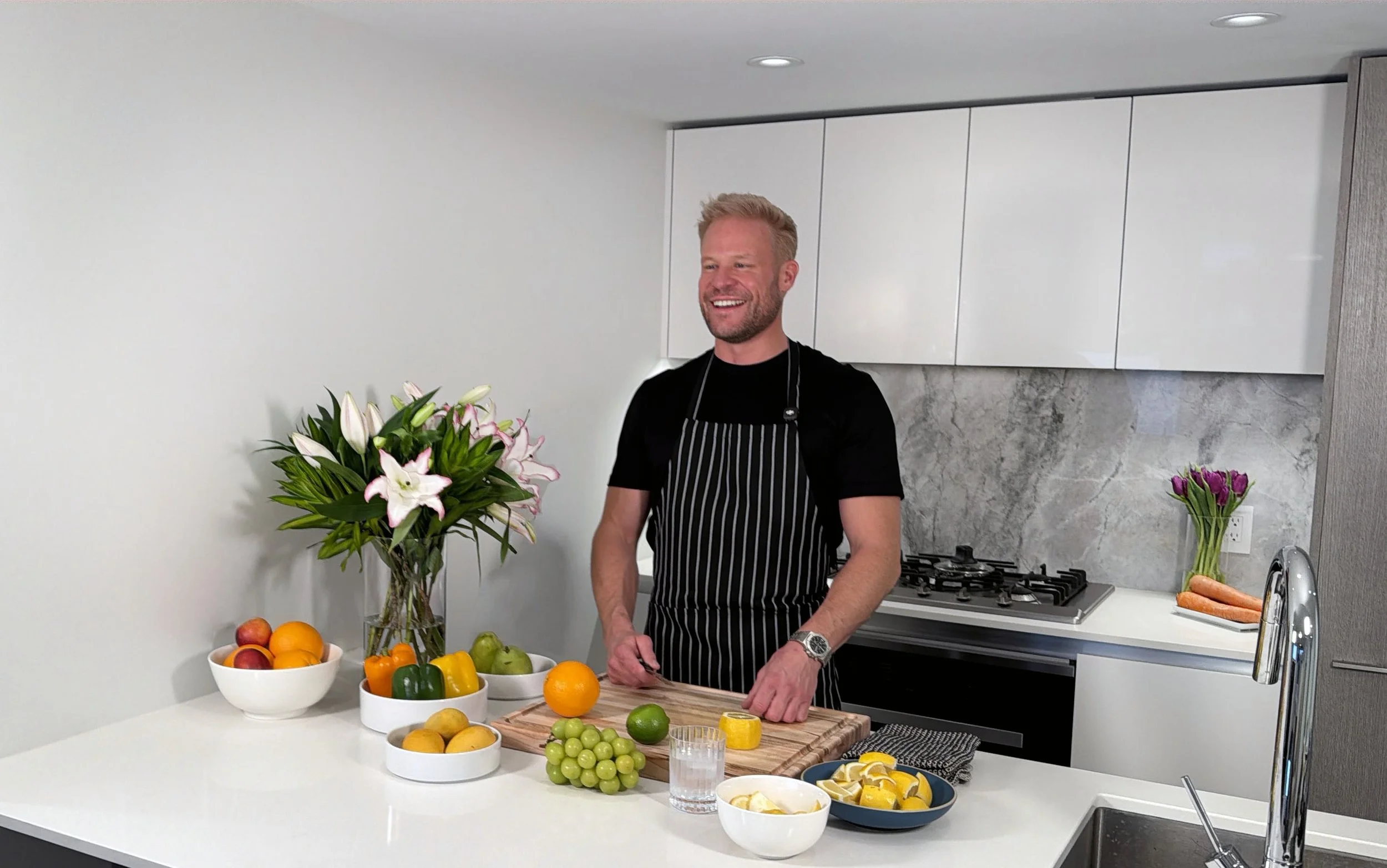 The founder of Financial Yoga, Ryan J. Jennings, stands in his kitchen wearing a black t-shirt and striped apron, slicing fresh citrus fruits.