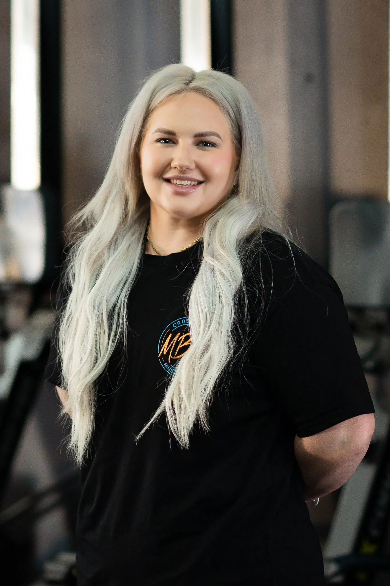 A smiling woman with long platinum blonde hair, wearing a black T-shirt with a logo, standing in an indoor setting with gym equipment in the background.