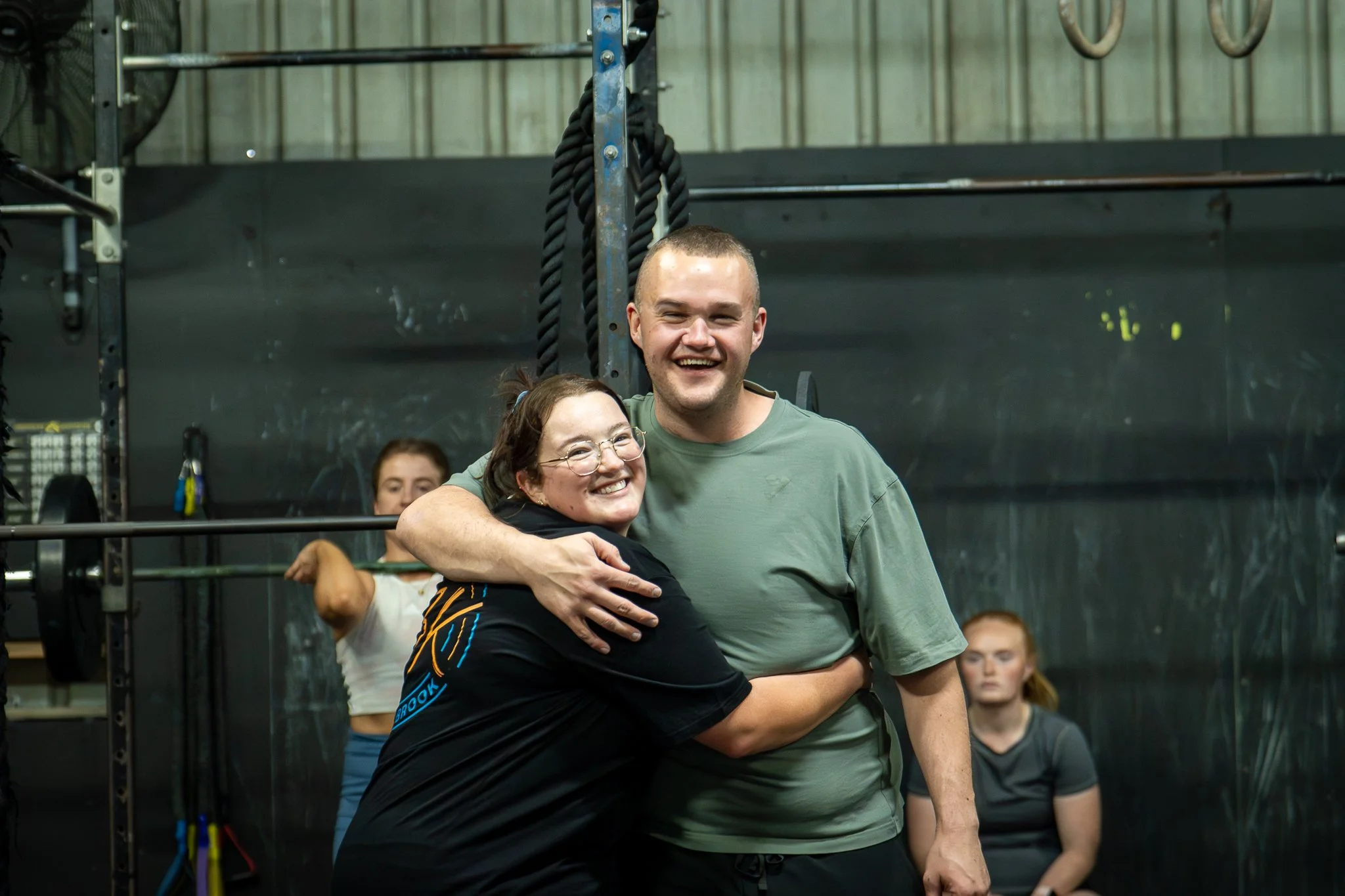 Two smiling people hugging in a gym with weights and ropes in the background.