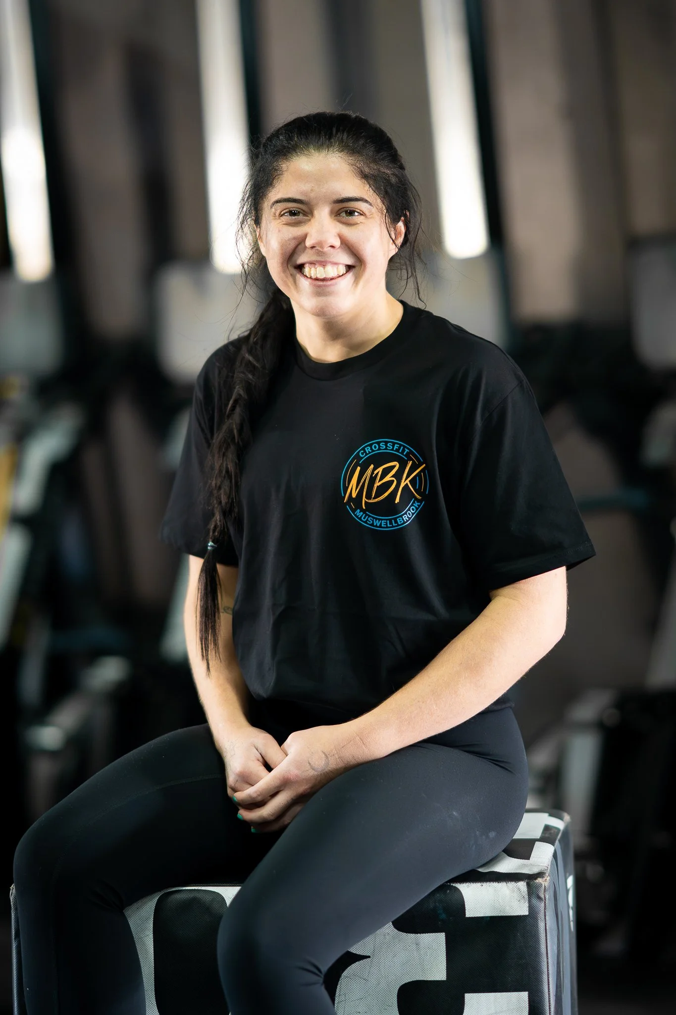 A woman with dark hair in a braid, smiling, sitting on a black and white box in a gym. She wears a black t-shirt with a logo that reads 'CrossFit Muswellbrook' and 'MBK'.