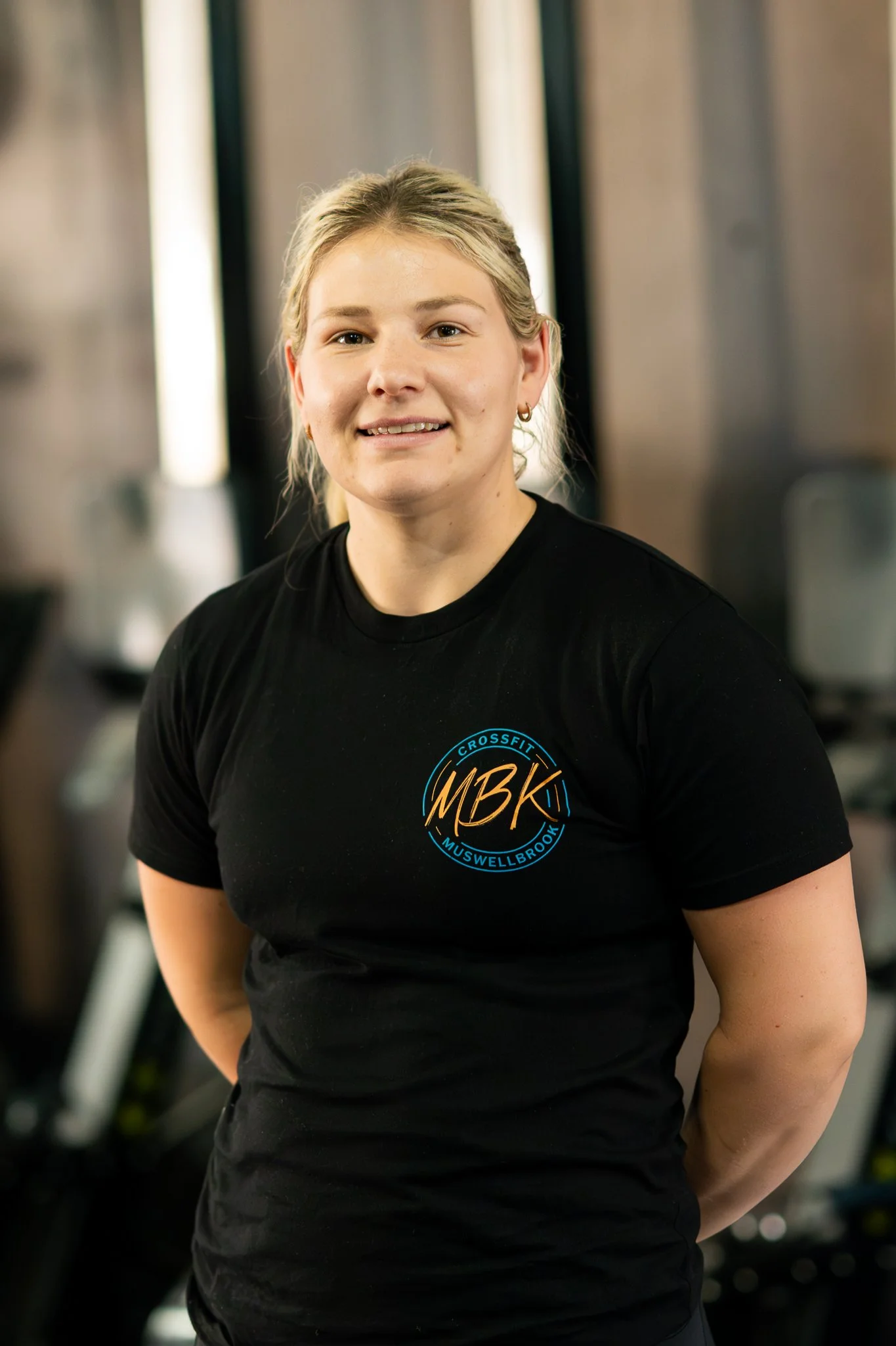 A young woman with blonde hair pulled back, wearing a black fitness shirt with the logo 'MBK Crossfit Muswellbrook,' stands in a gym with fitness equipment in the background.