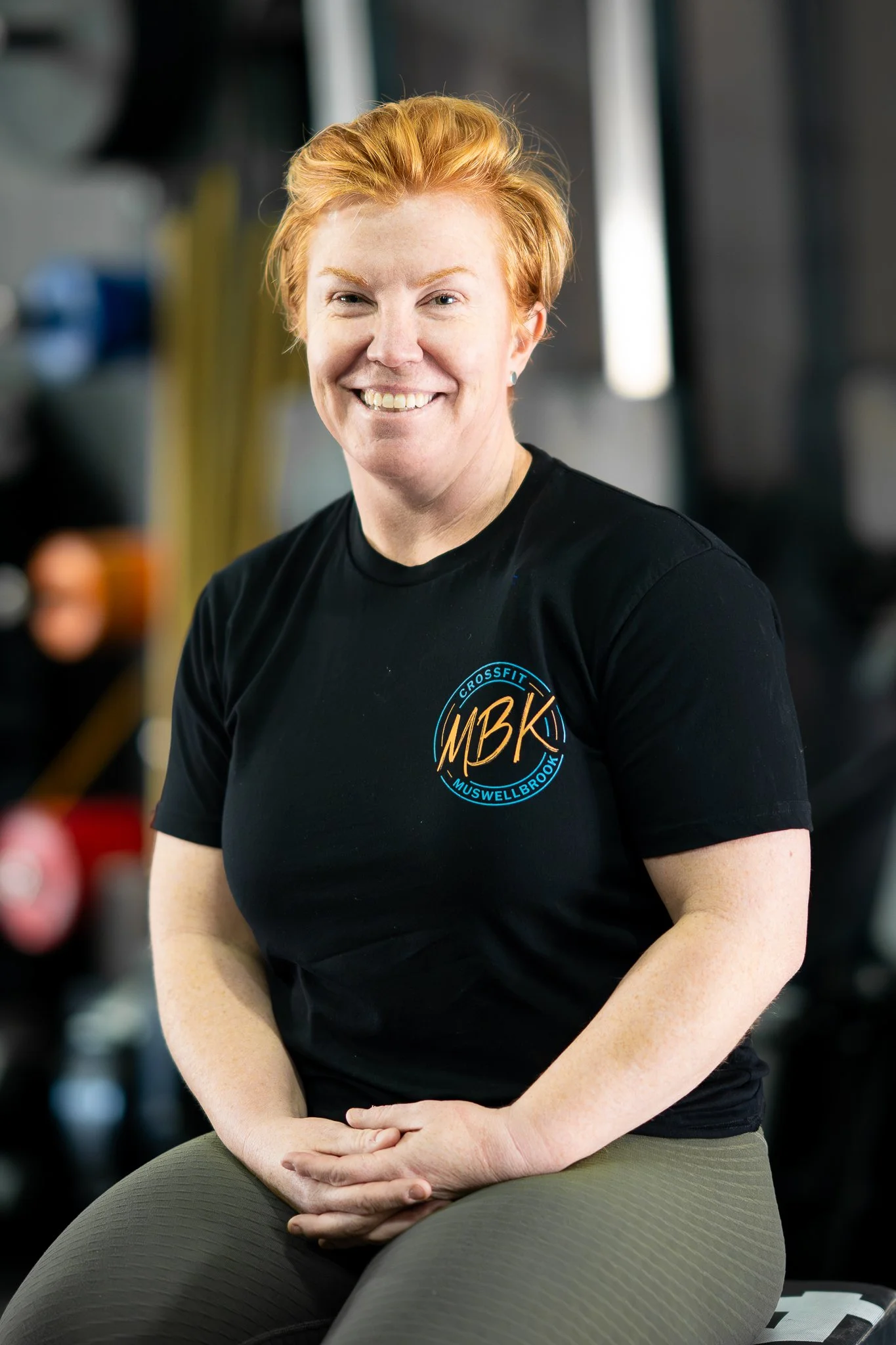 A woman with short red hair smiling at the camera, wearing a black CrossFit shirt with a logo on the chest, sitting in a gym setting.