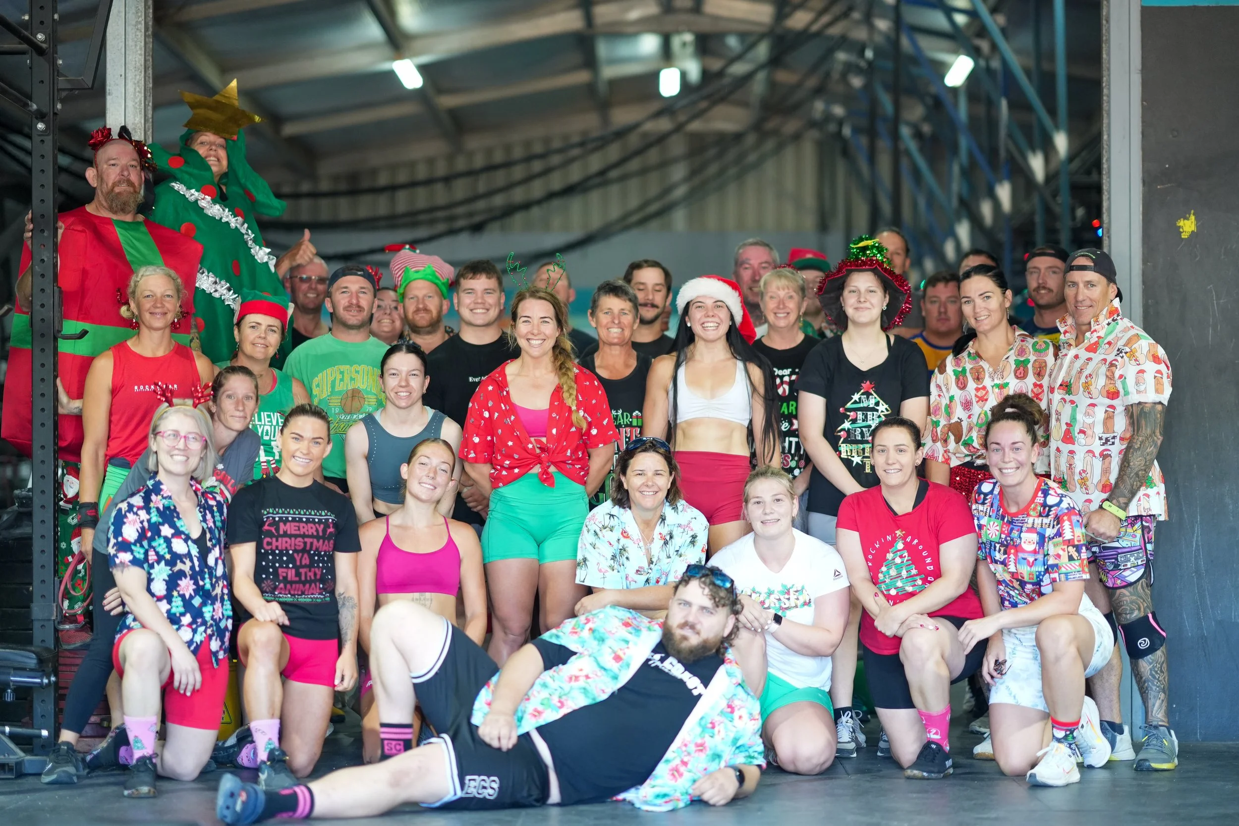 Group of people dressed in Christmas-themed attire including Santa hats, elf costumes, and holiday shirts, posing for a photo indoors with festive accessories.