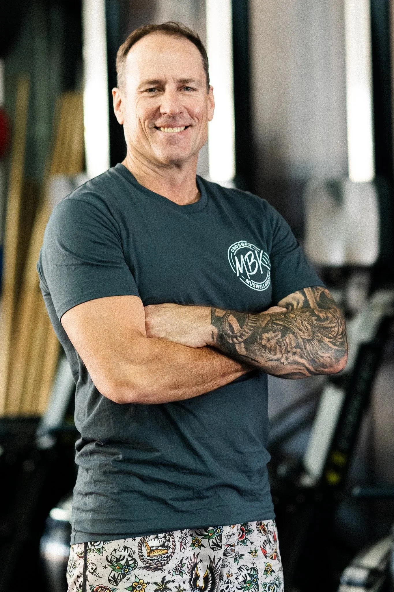 A man with short brown hair and tattoos on his right arm stands with arms crossed, smiling, in a gym.
