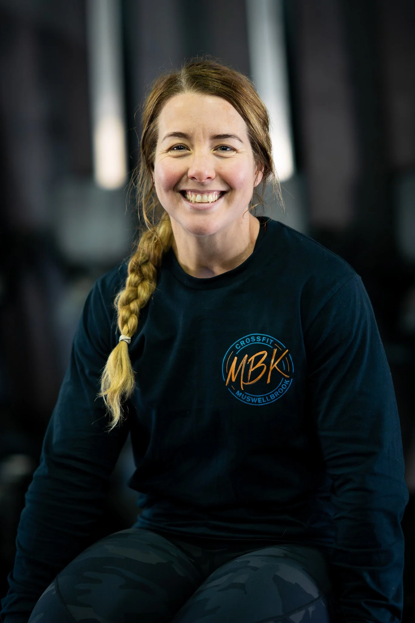A woman with red hair in a braid, smiling, wearing a black long-sleeve shirt with a CrossFit logo that reads 'MBK Muswellbrook' on it, in a gym.