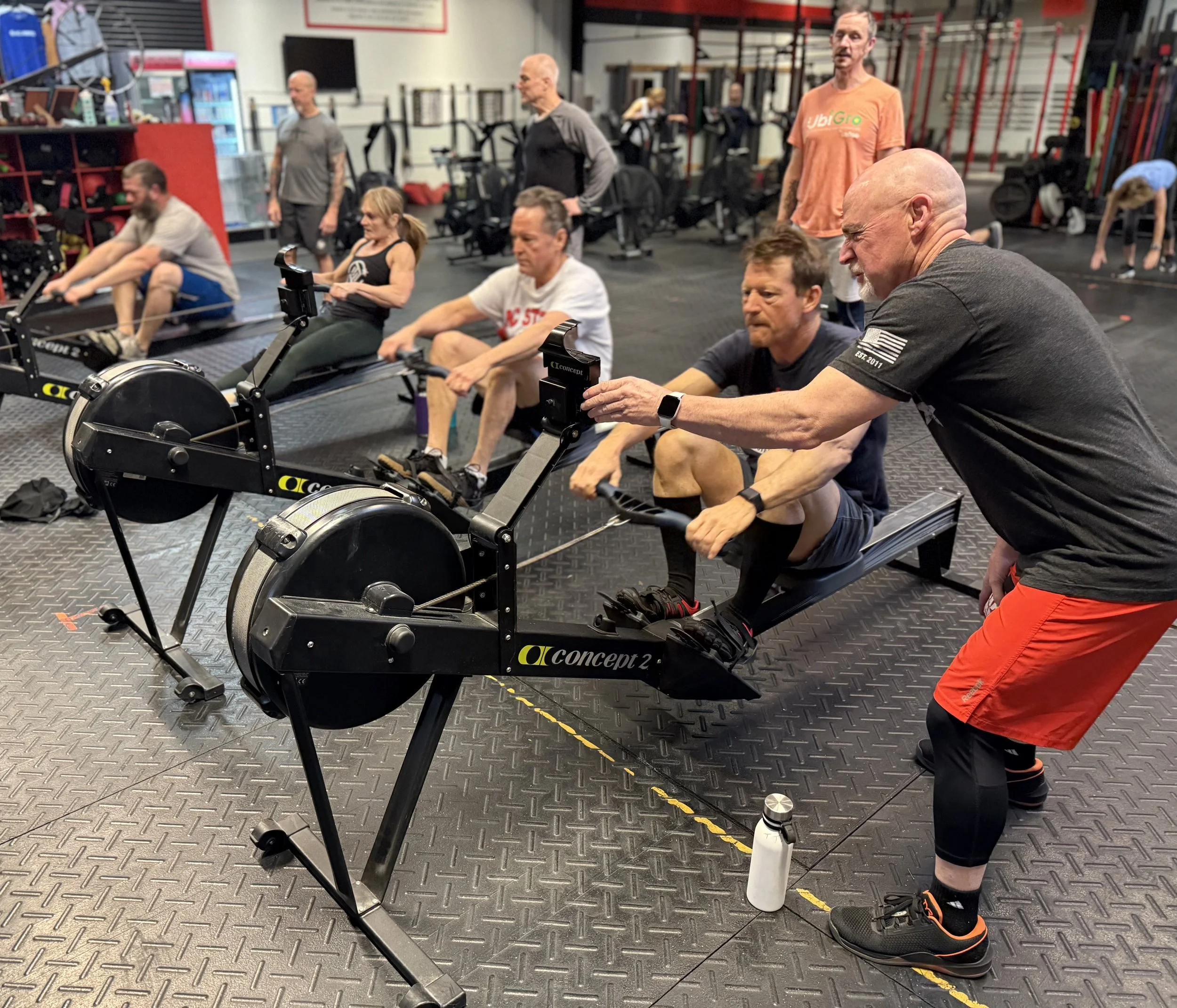 Several Adult men and women of different ages use rowing machines side by side, during a group workout at Crossfit Full Potential. One partner in the foreground is reading the time on a rower's display as he prepares to take over the machine.
