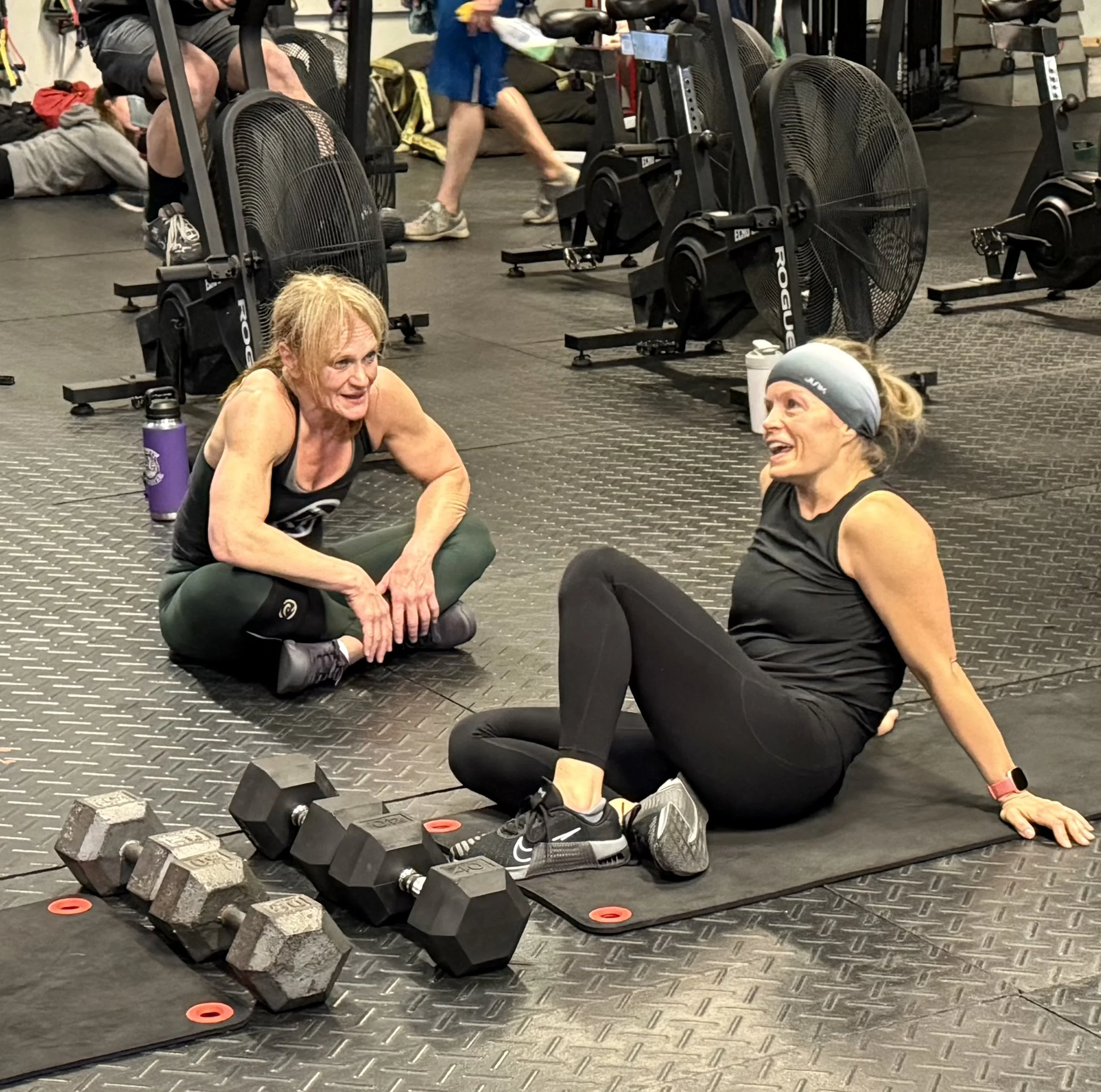 Two women sit on the floor of the gym at CrossFit Full Potential, talking as they rest. One woman is sitting with her legs criss-crossed as she listens to the other talk, as she reclines slightly on a mat in front of several large dumbbells.