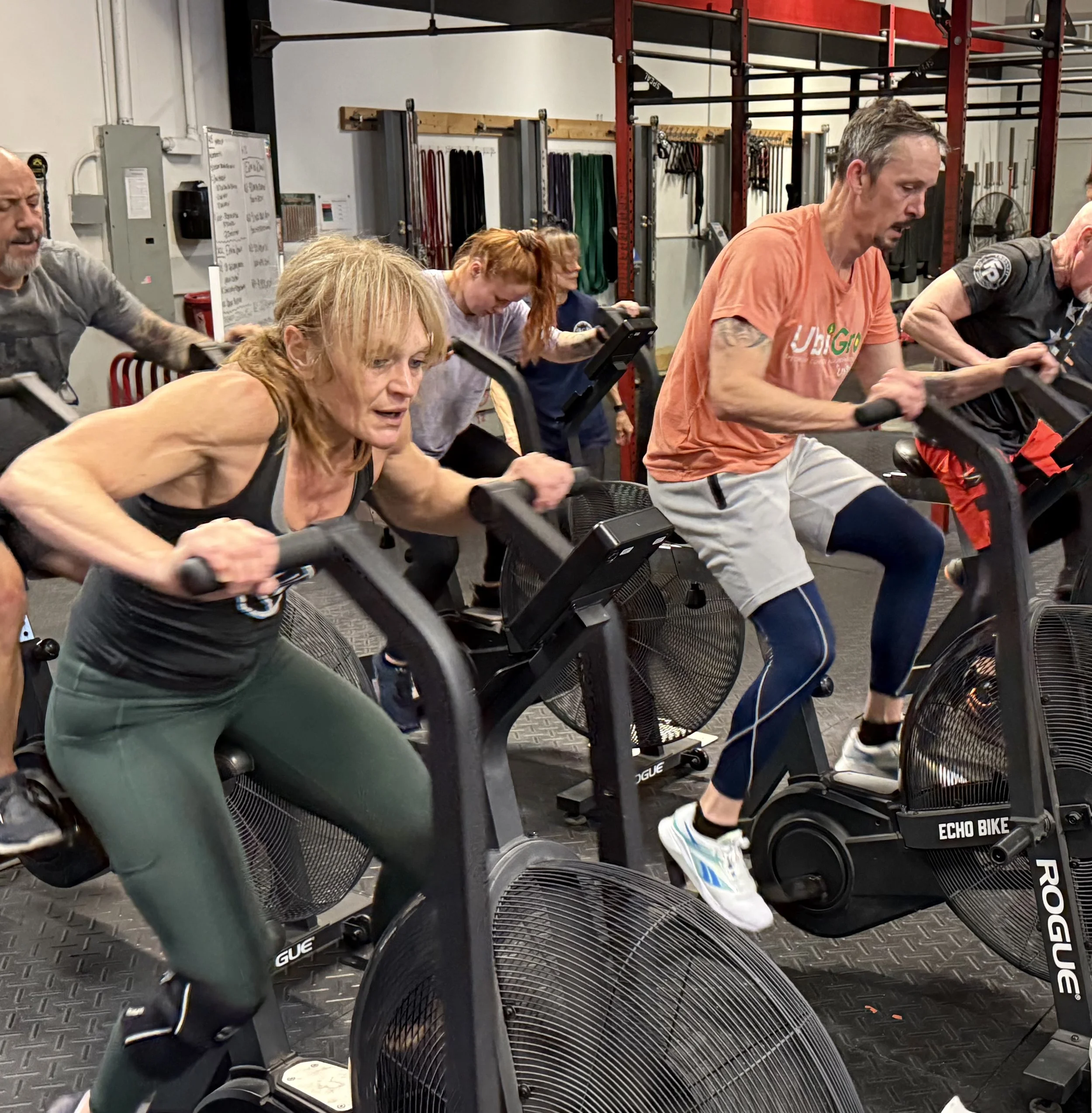 Men and women of various ages pedal air bikes in a CrossFit gym during a group workout. The bikes are in two rows side by side, and each athlete is leaning forward with effort.