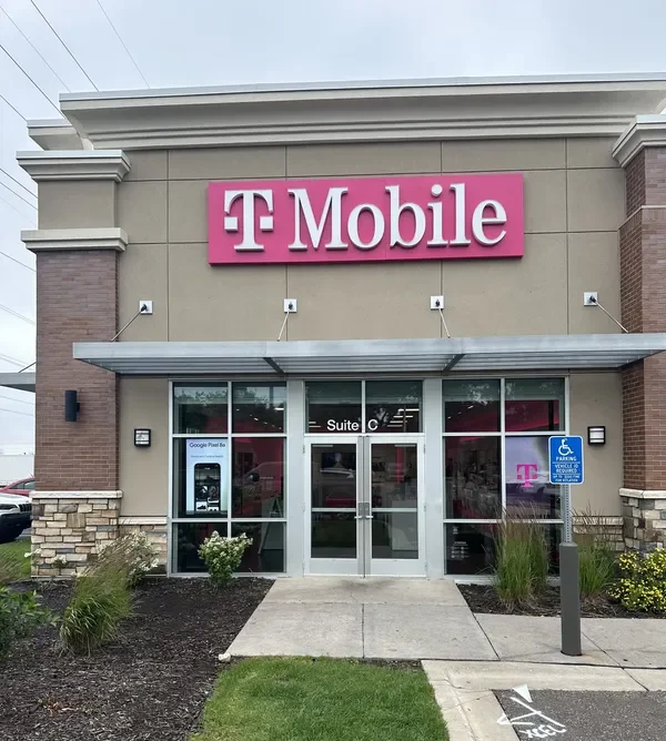 Exterior of a T-Mobile store with a pink sign, glass doors, a wheelchair accessible parking sign, and a small landscaped area in front. HVAC Services New York City, USA.